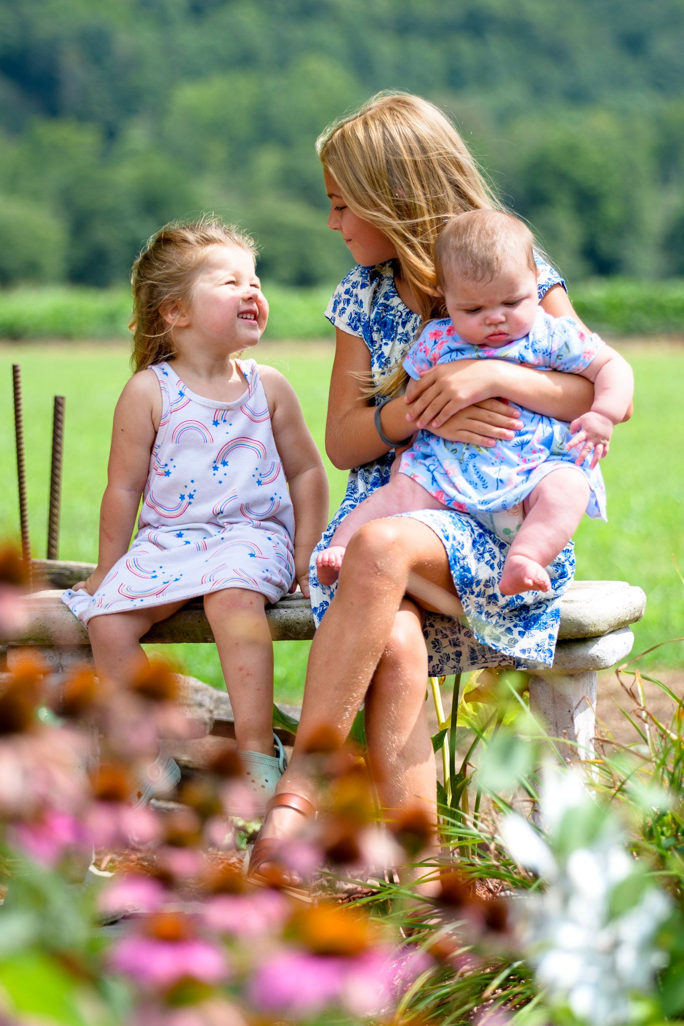 A mother in a floral dress sits on a stone bench holding a baby while her young daughter laughs beside her.