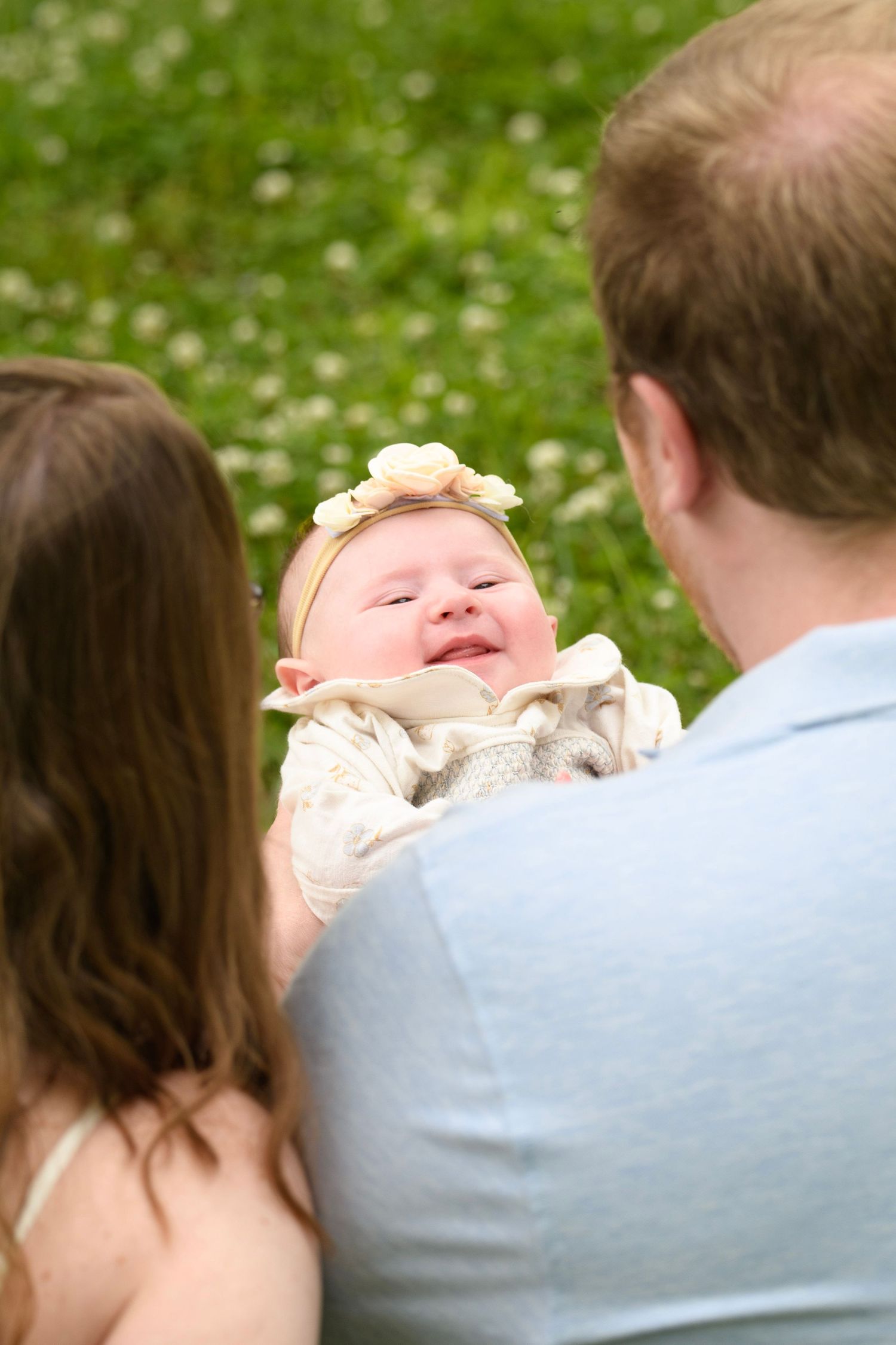 Smiling newborn baby with flower headband being held by parents on a grass field.