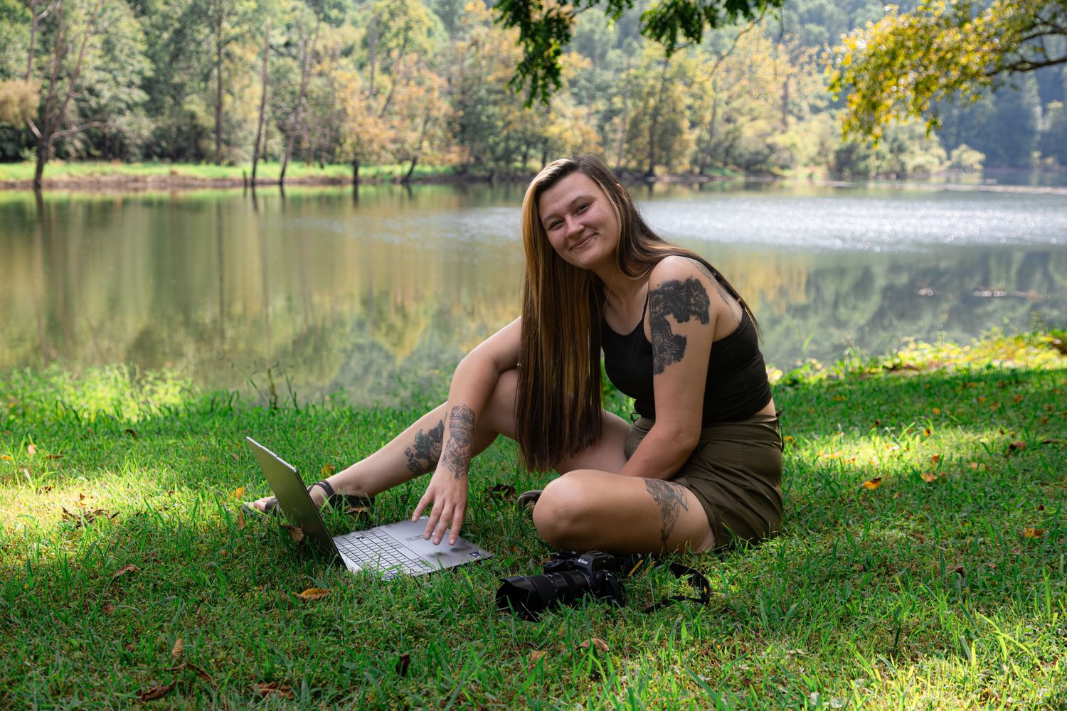 Young woman with tattoo sitting on grass by a lake, smiling while using a laptop outdoors in a park.