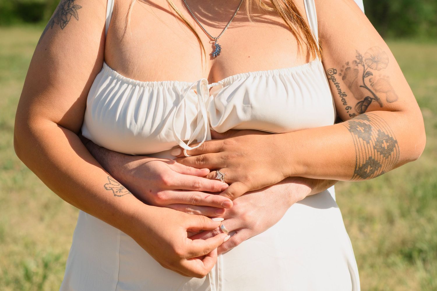 Close-up of two women in white dresses holding hands showing tattoos in sunny field.