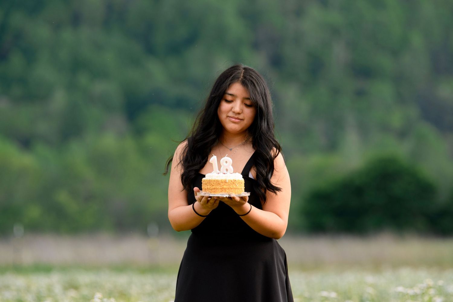Young woman in black dress holding a birthday cake with candles in a wildflower meadow.