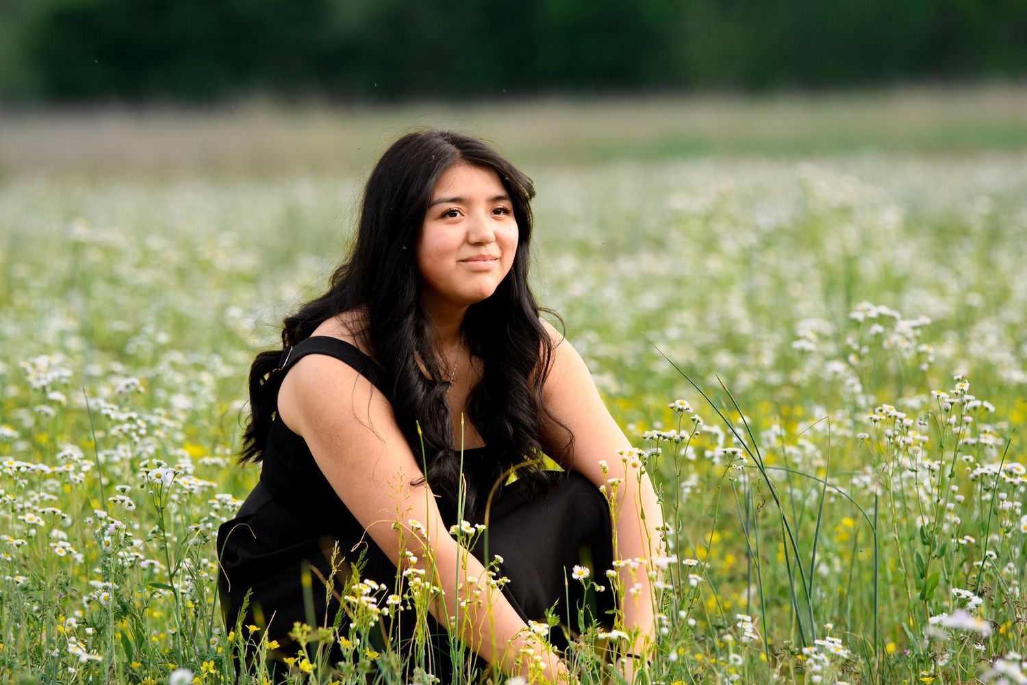 Young woman with long dark hair sits smiling in a wildflower meadow wearing a black outfit.