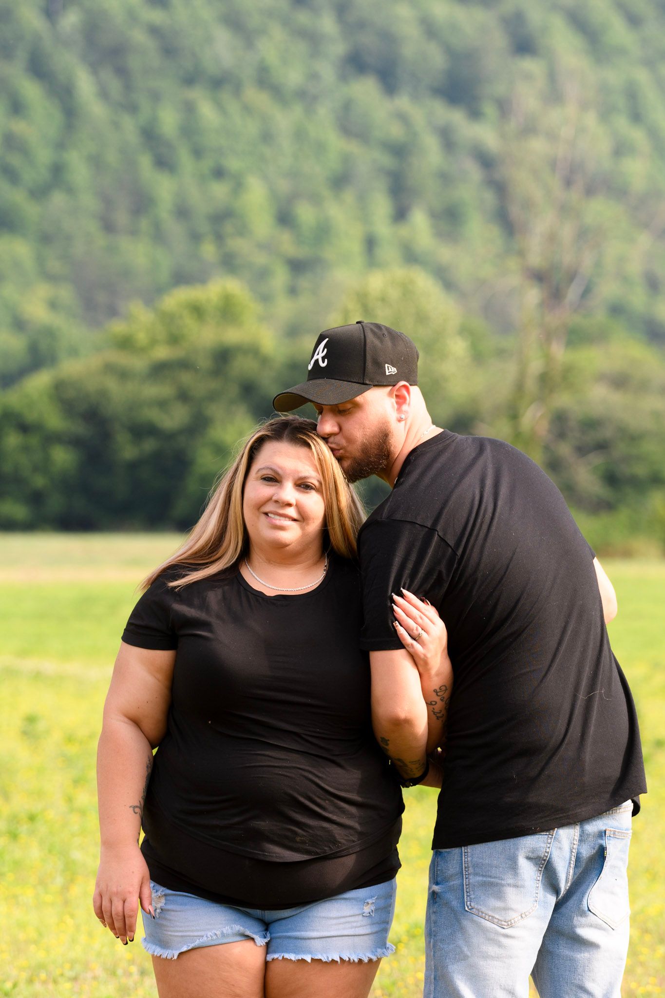 Couple in matching black shirts posing outdoors with lush green hills, man kissing woman's head in a field.