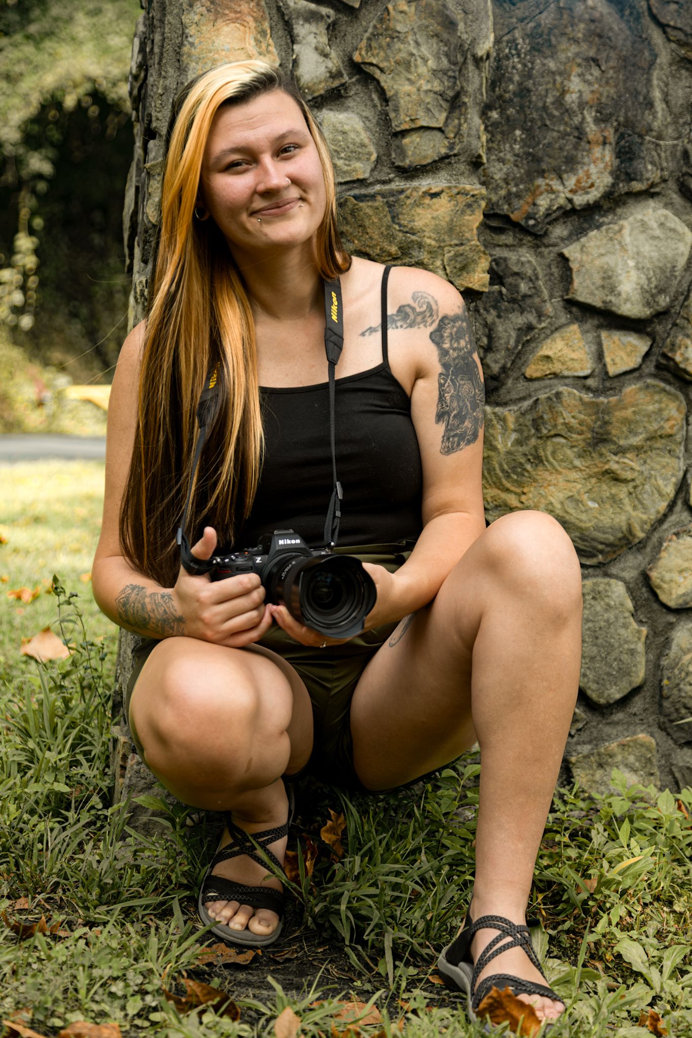 Young woman with long hair and tattoos crouches against stone wall holding a DSLR camera outdoors.