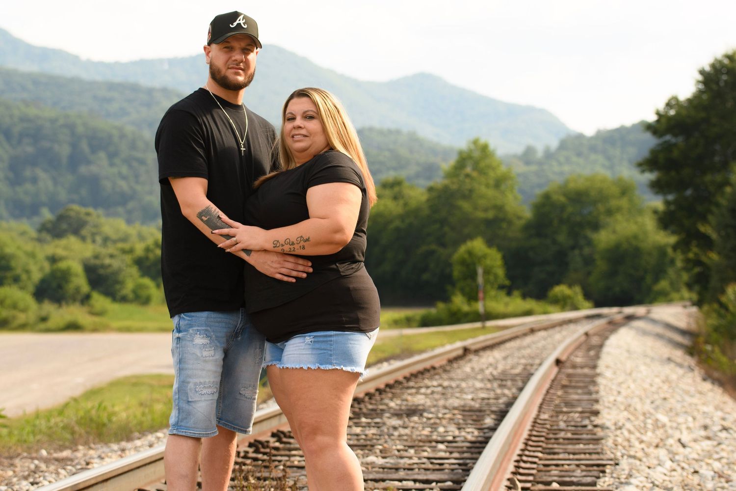 Couple posing on railroad tracks with scenic mountain backdrop on a sunny day.