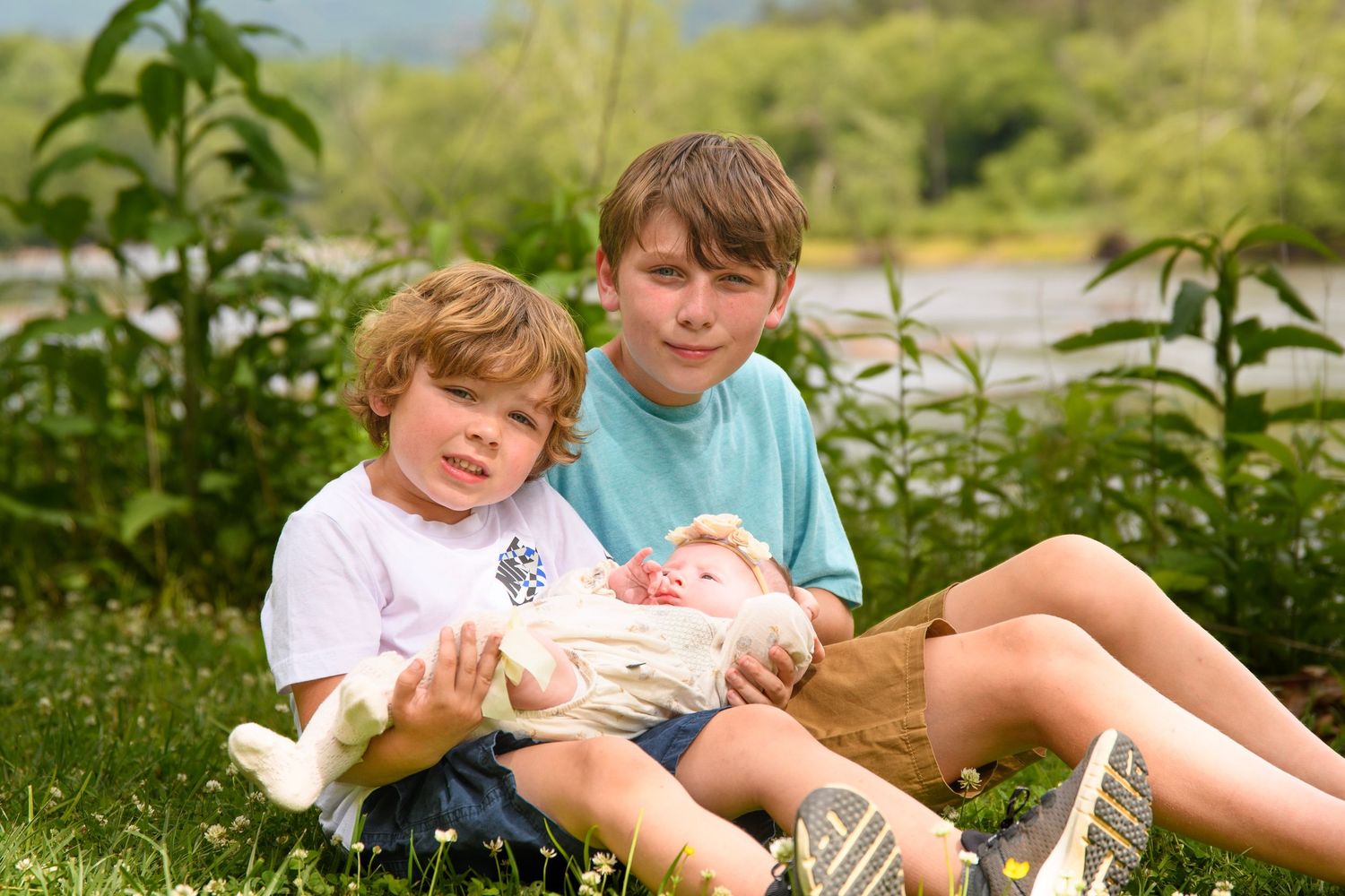 Two young brothers sitting in grass near a river, older boy holding a newborn baby.