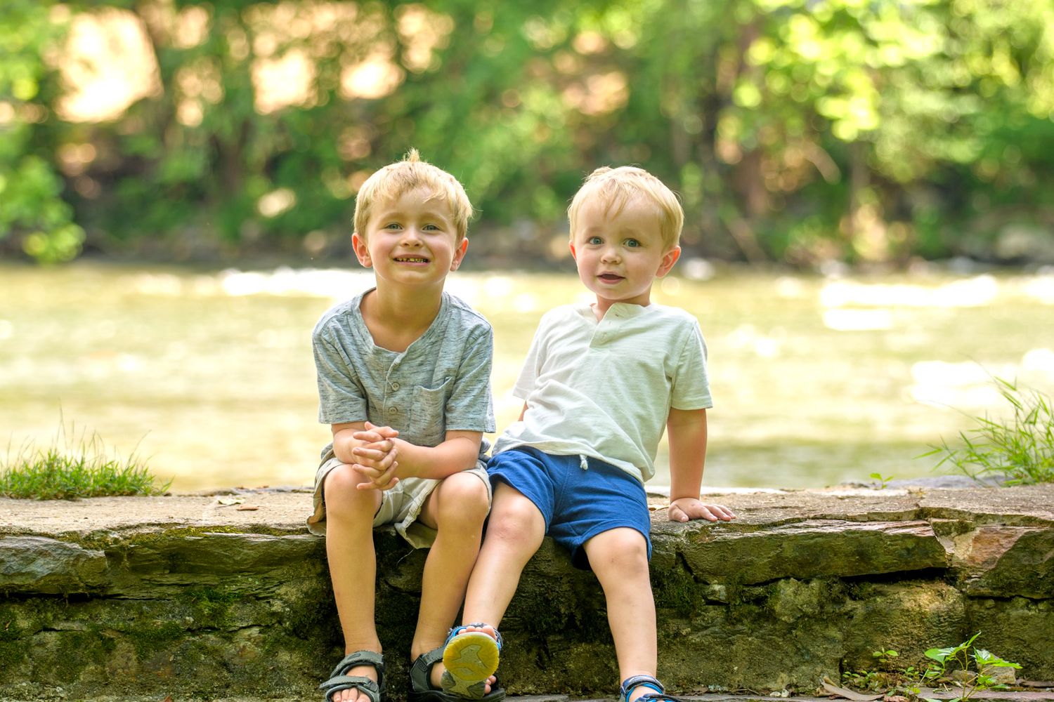 Two young blonde brothers sit together on a stone ledge near a river, smiling at the camera outdoors.