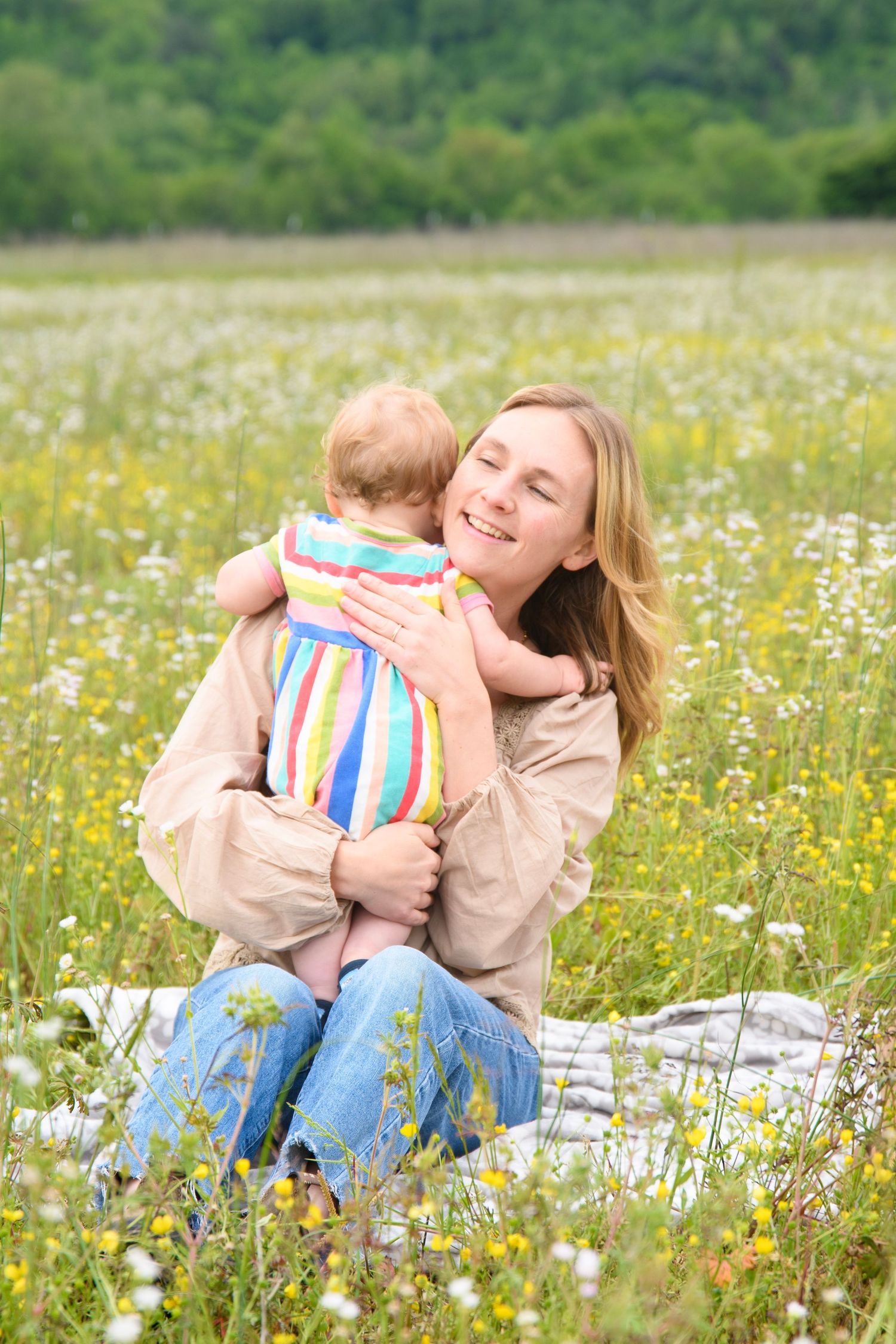 Smiling mother hugging baby in rainbow striped outfit while sitting in a blooming wildflower meadow.