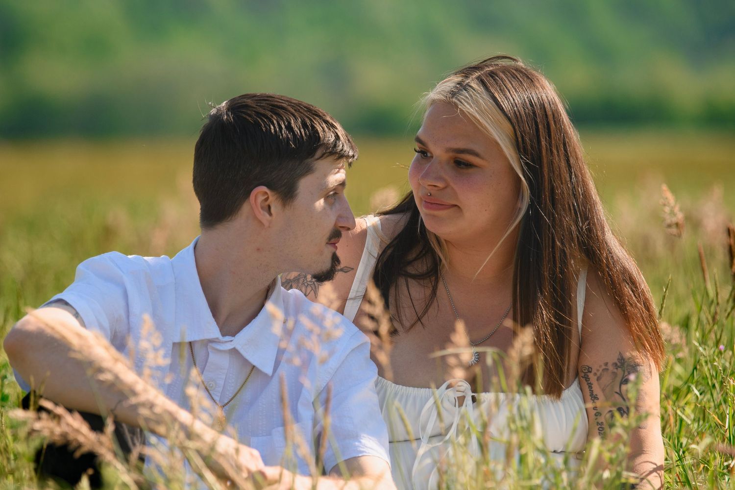 Young couple sitting together in a sunny green field, gazing at each other romantically.