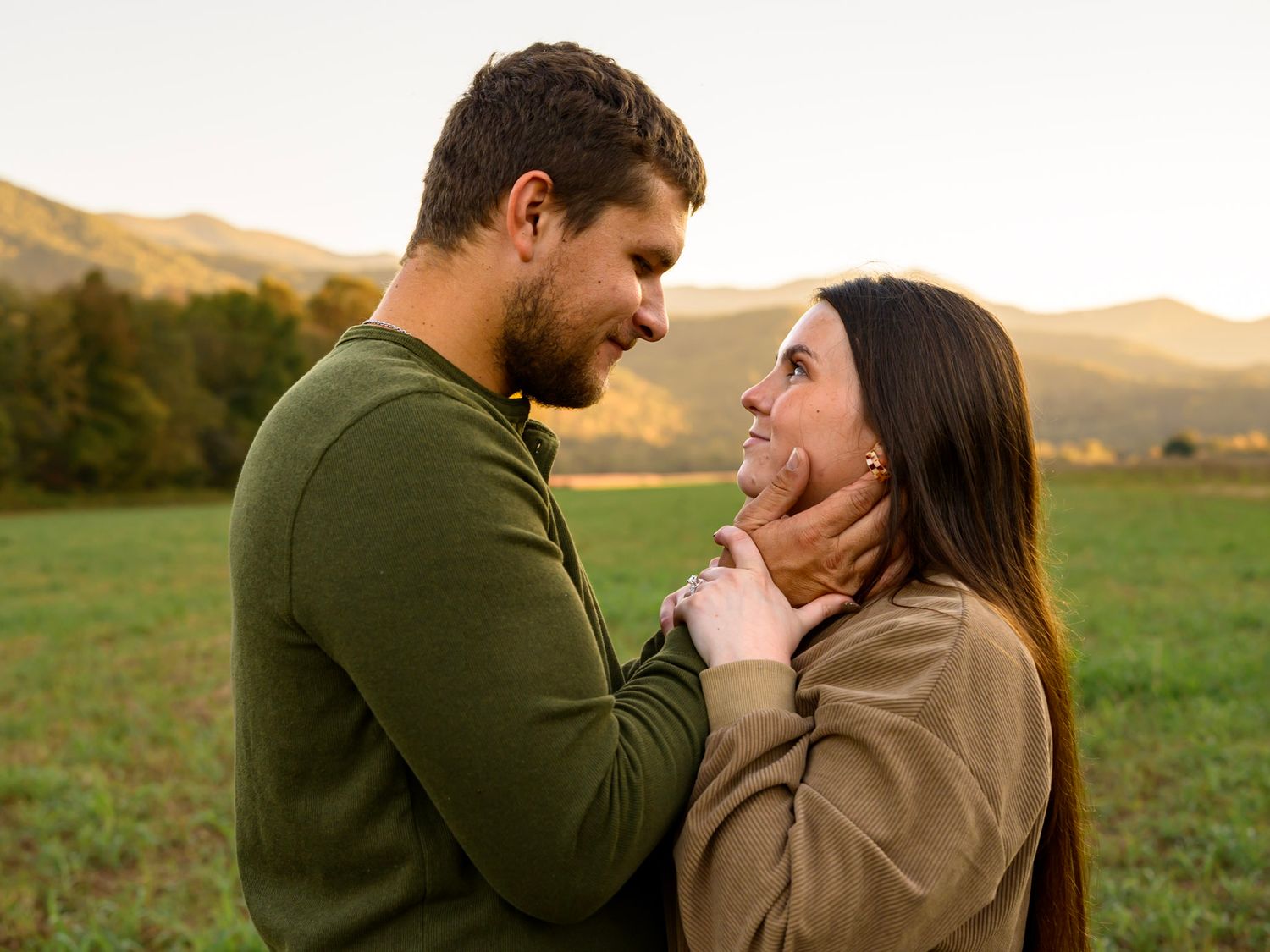 Smiling couple shares a tender moment in a green meadow at sunset, with the woman cradling the bearded man's face and mountains behind them.