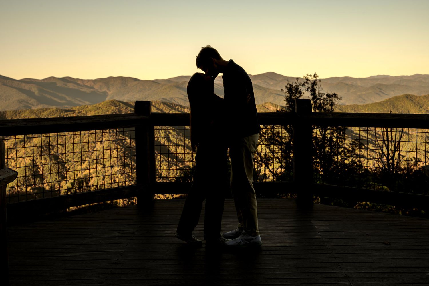 Silhouette of a couple kissing on a wooden deck at sunset with a railing and layered mountain ridges glowing in the background.