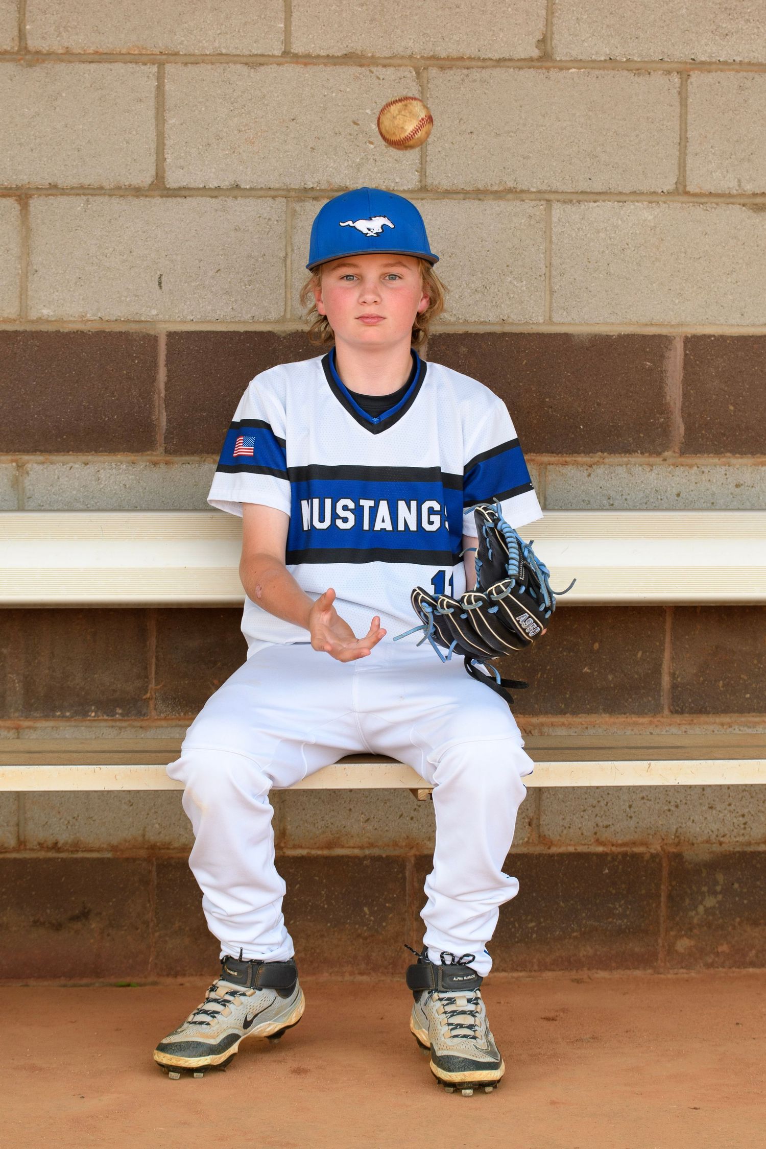 Young baseball player in Mustangs uniform sitting in dugout tossing a ball, wearing blue cap and glove.