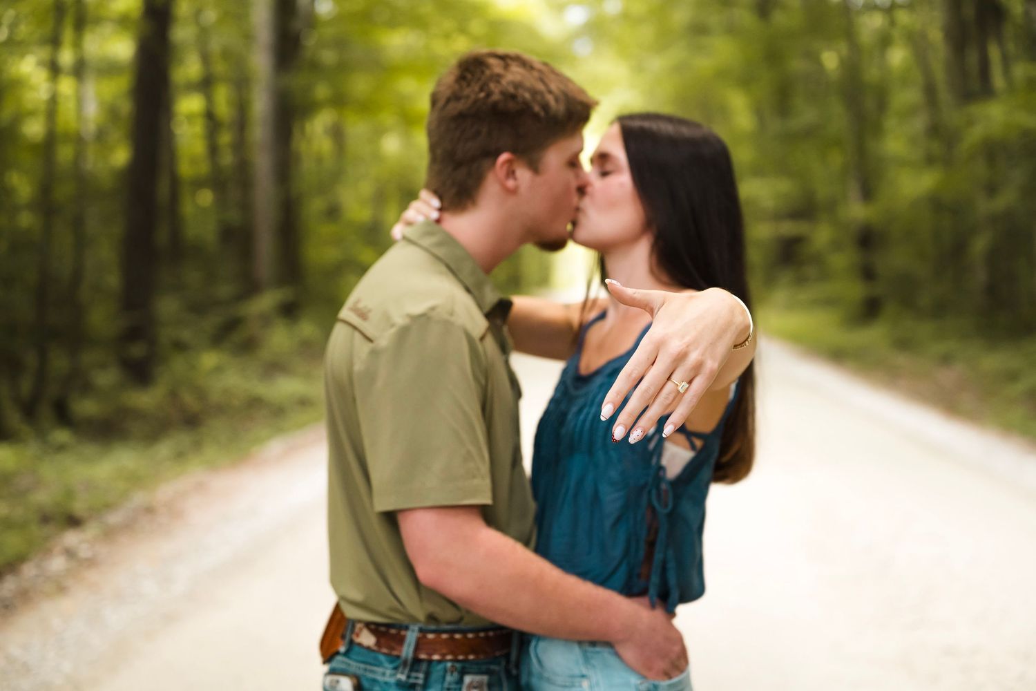 Couple sharing a romantic kiss on a tree-lined dirt road, woman displaying engagement ring outdoors.
