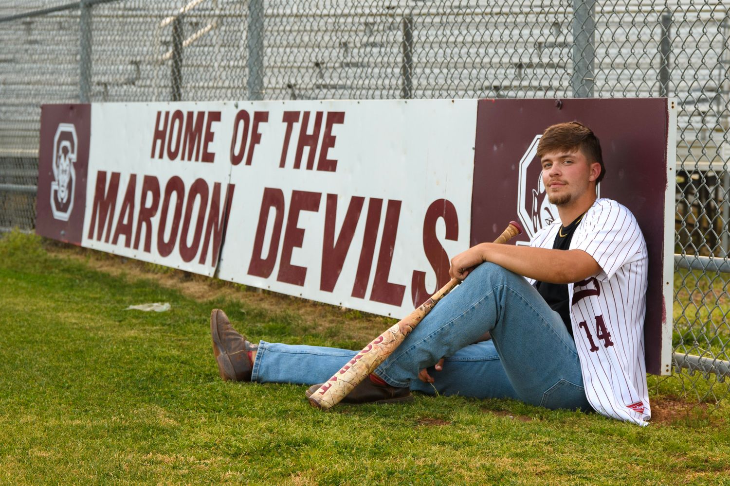 Teen baseball player in jersey sits beside a Maroon Devils sign holding a bat for a sports senior portrait.