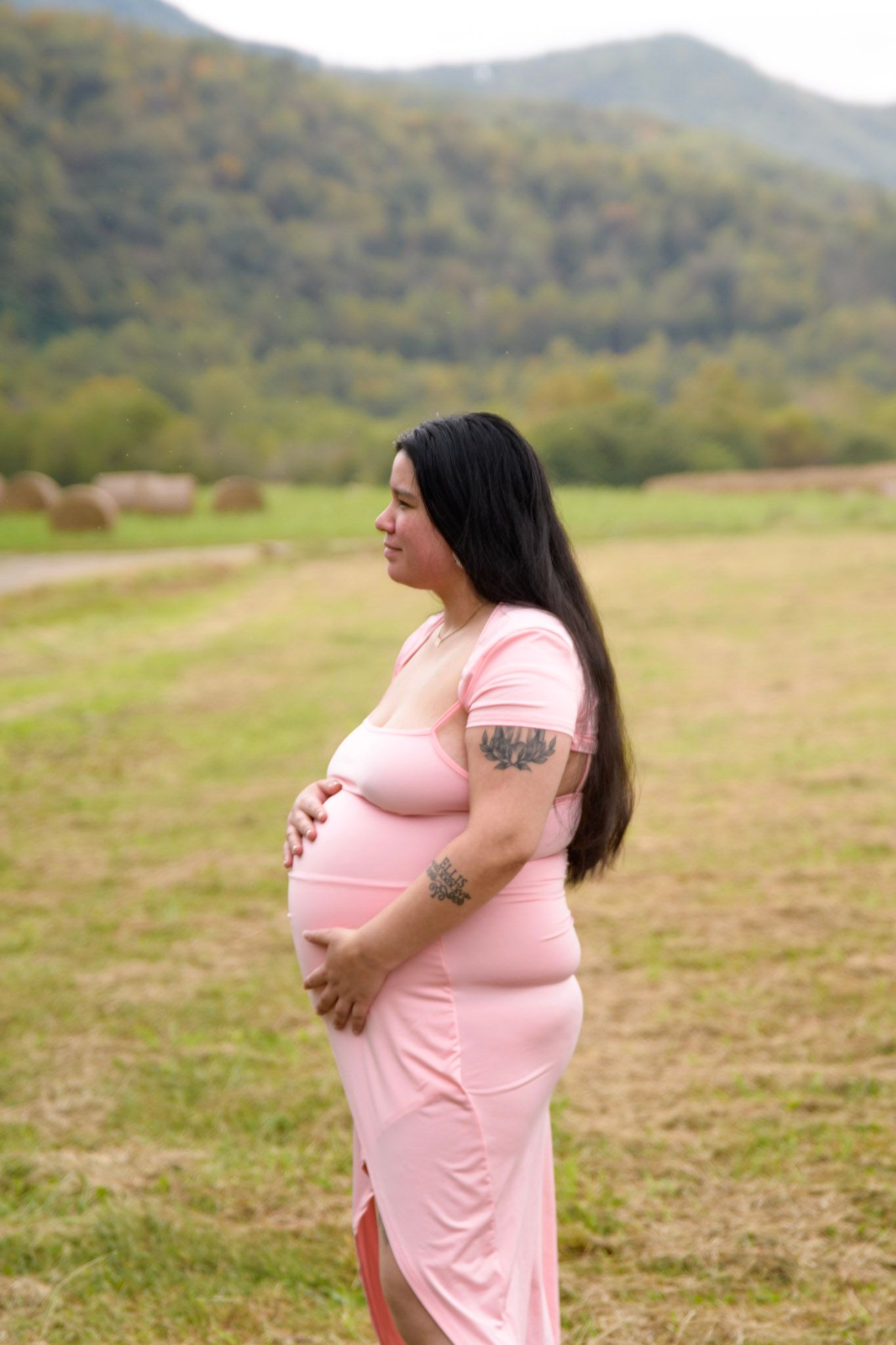 Pregnant woman in pink dress standing in field with scenic mountain backdrop.