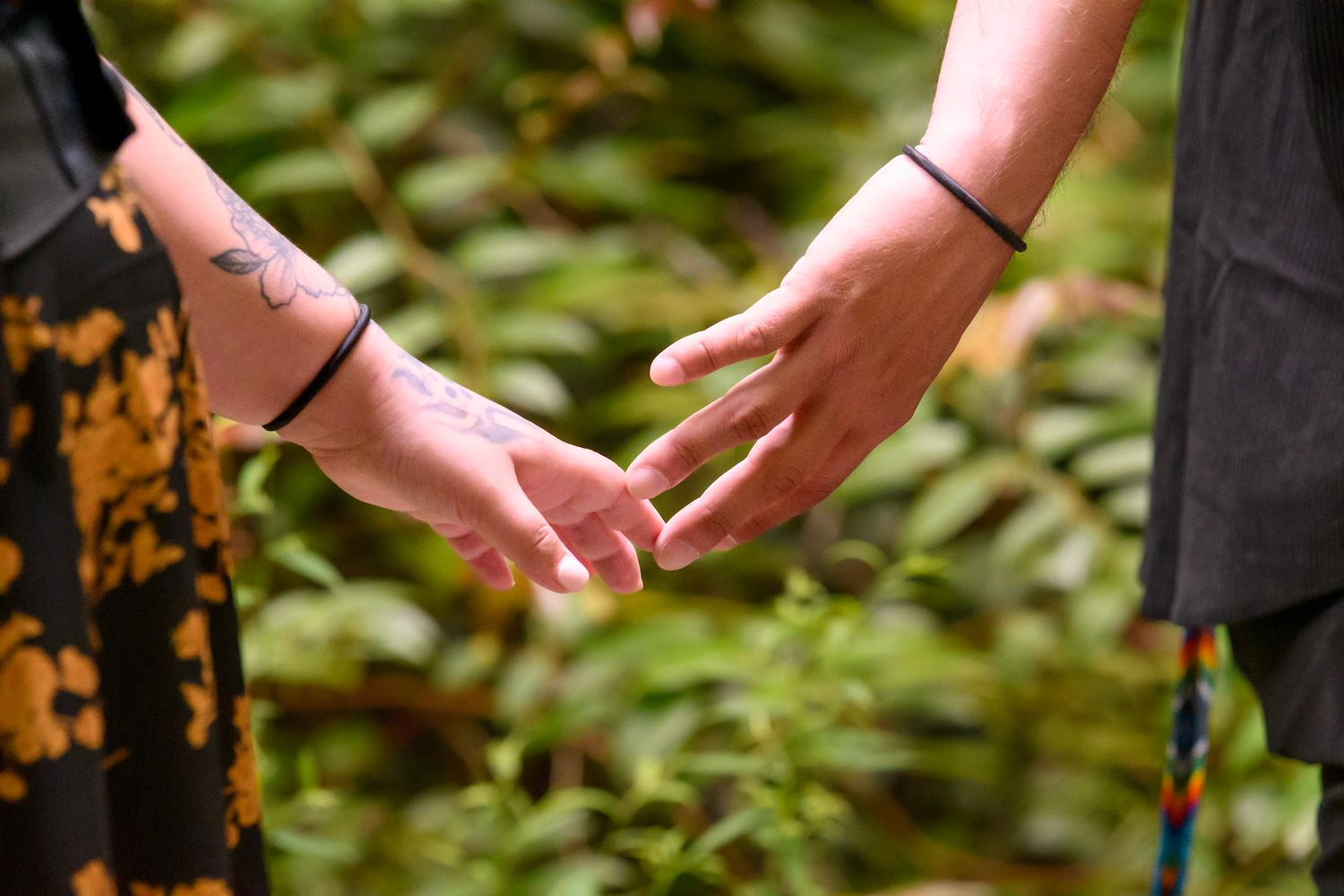 Two people with matching black bracelets gently touching hands outdoors with lush green foliage background.