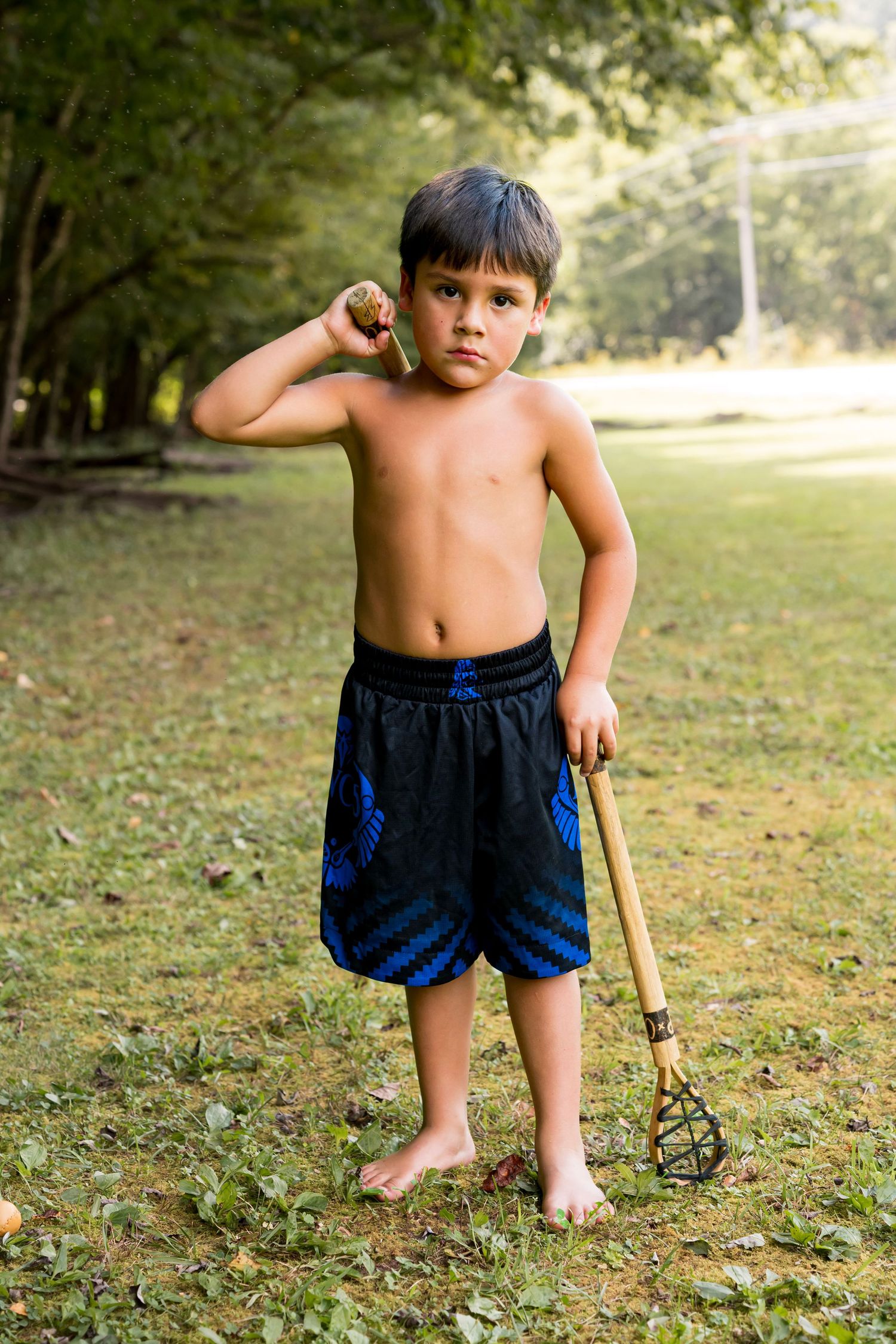 Shirtless boy holding a garden rake and a ball, standing barefoot on grass in a park.