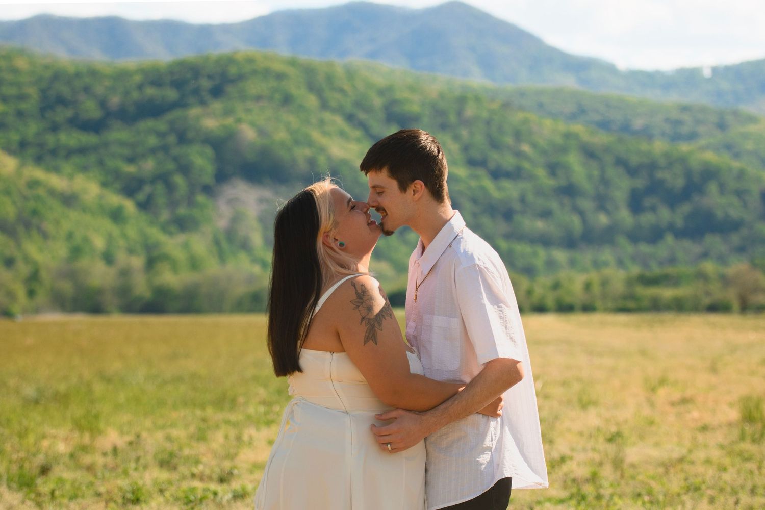 A couple shares a romantic kiss in a lush green field with mountain views in the background.