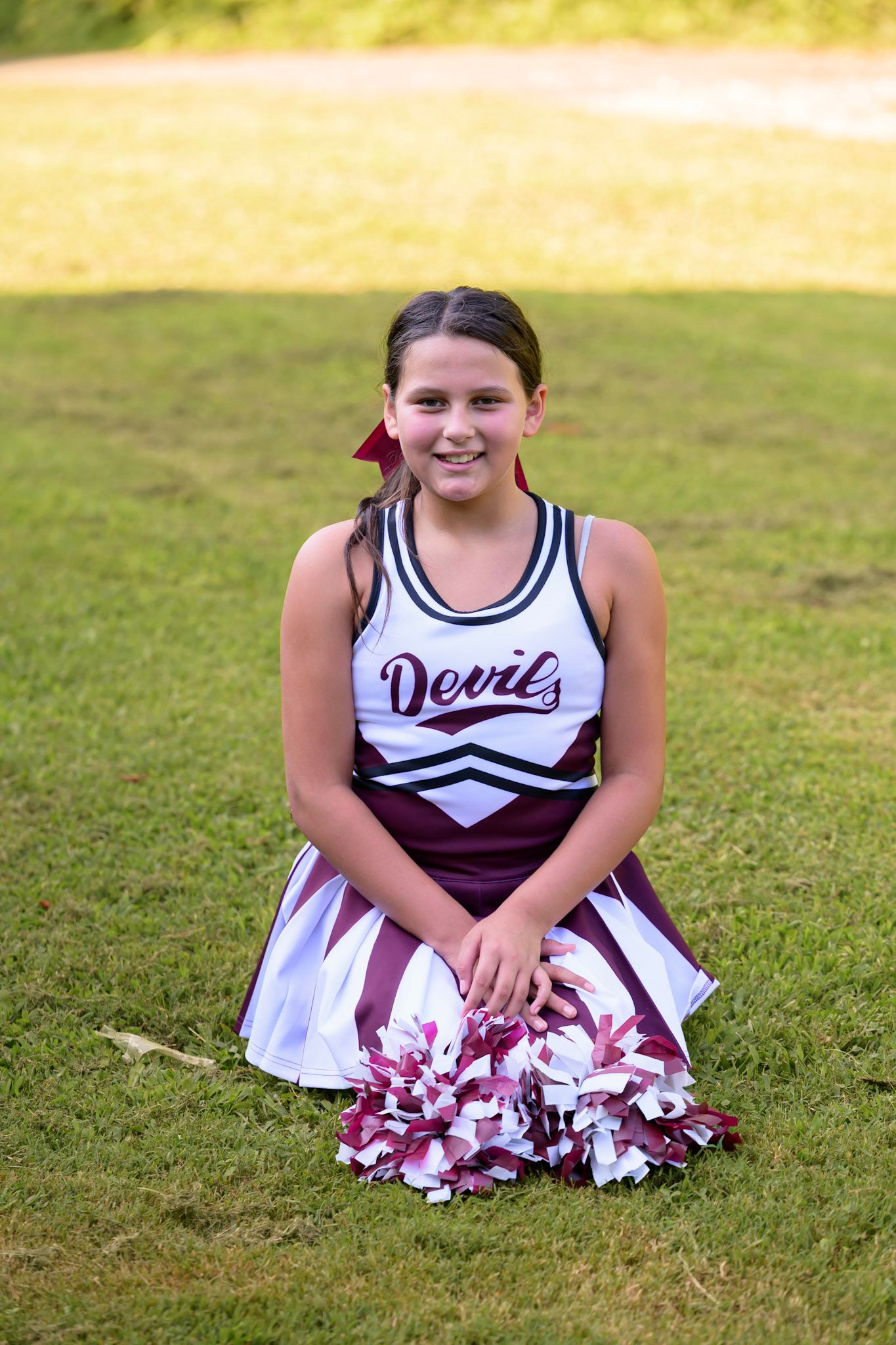 Young cheerleader in Devils uniform sitting on grass field holding maroon and white pom-poms, smiling for portrait photo.