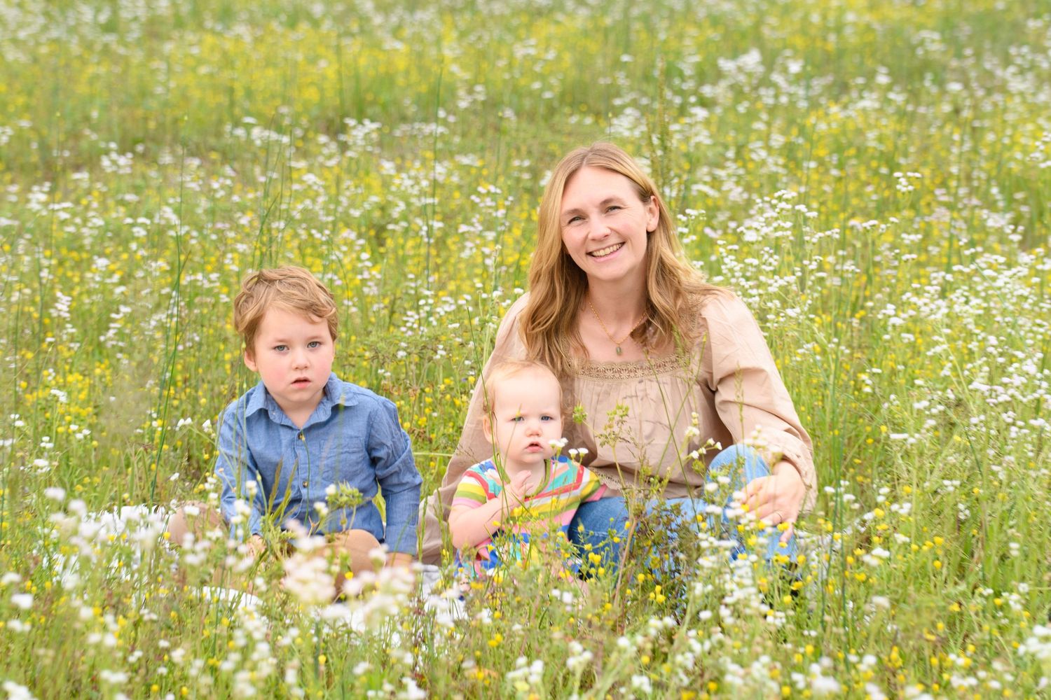 Smiling mother sitting with two young children in a sunny wildflower meadow field.