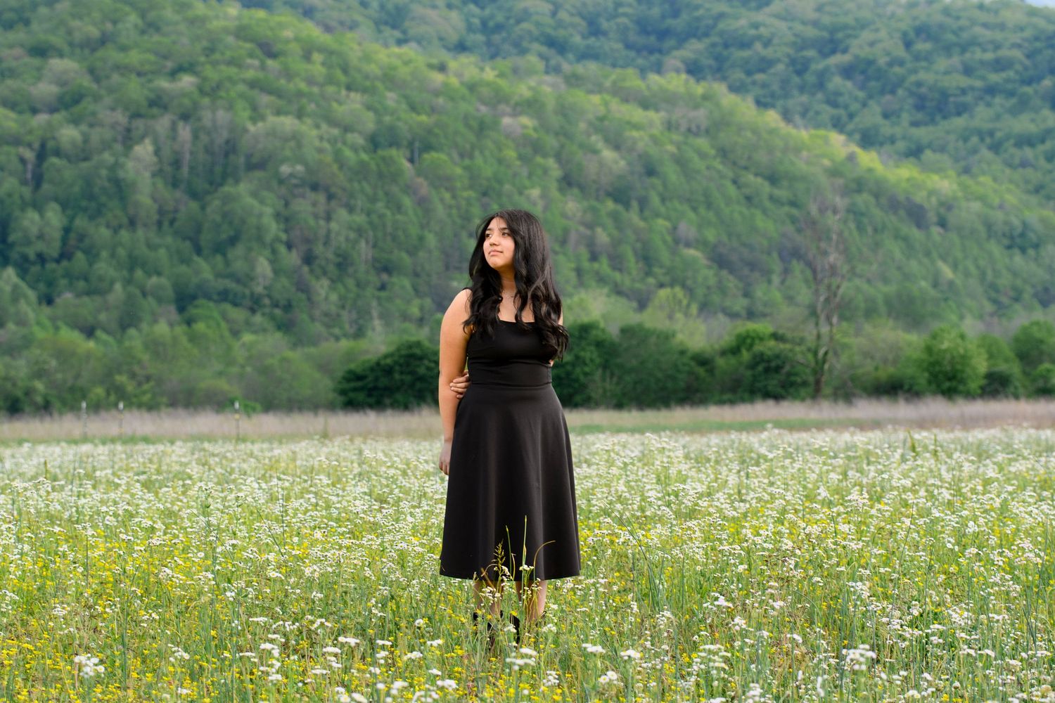 Young woman in black dress standing in white wildflower meadow with green mountain backdrop.