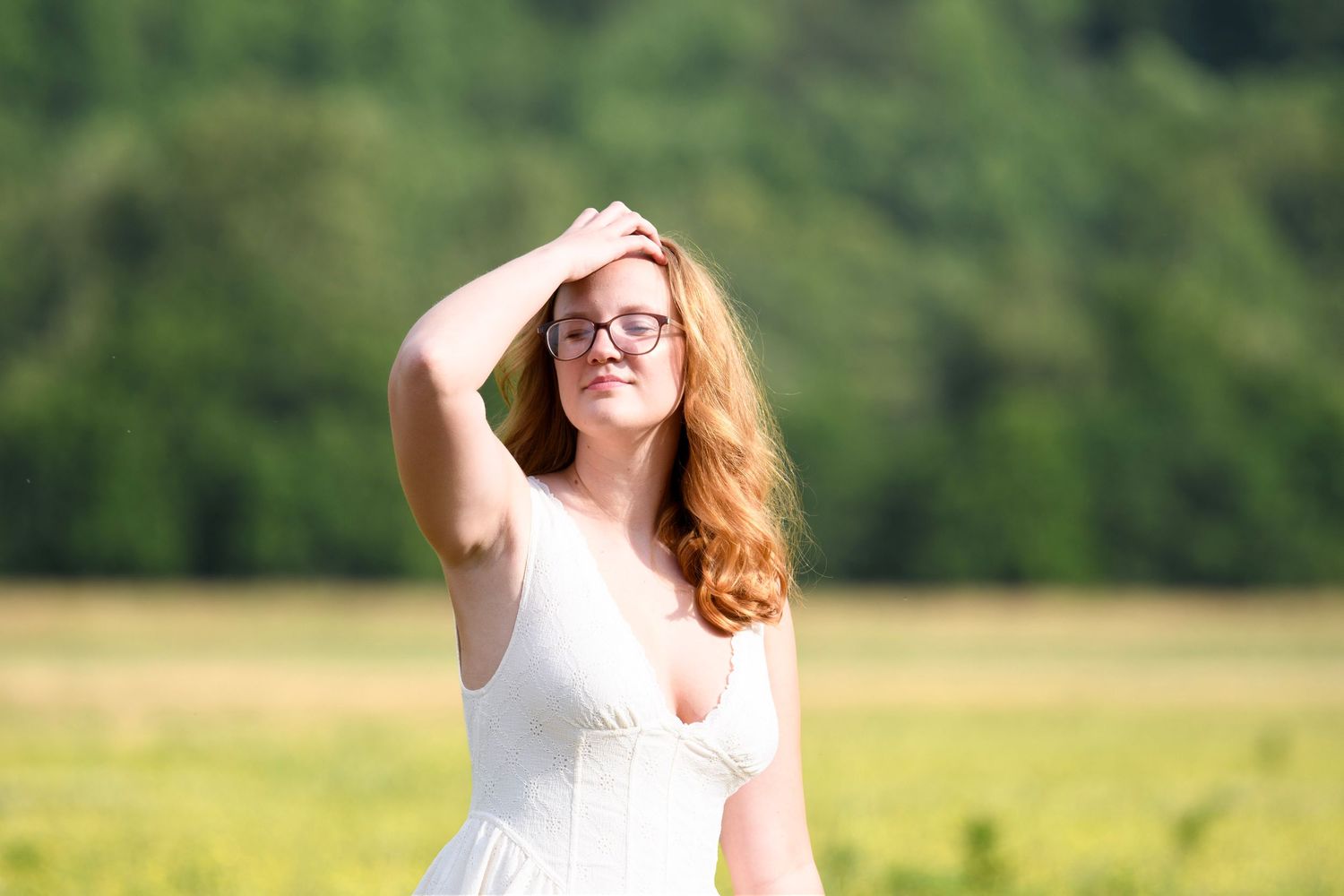 Young redhead woman in white dress and glasses poses in sunny green field with trees in background.