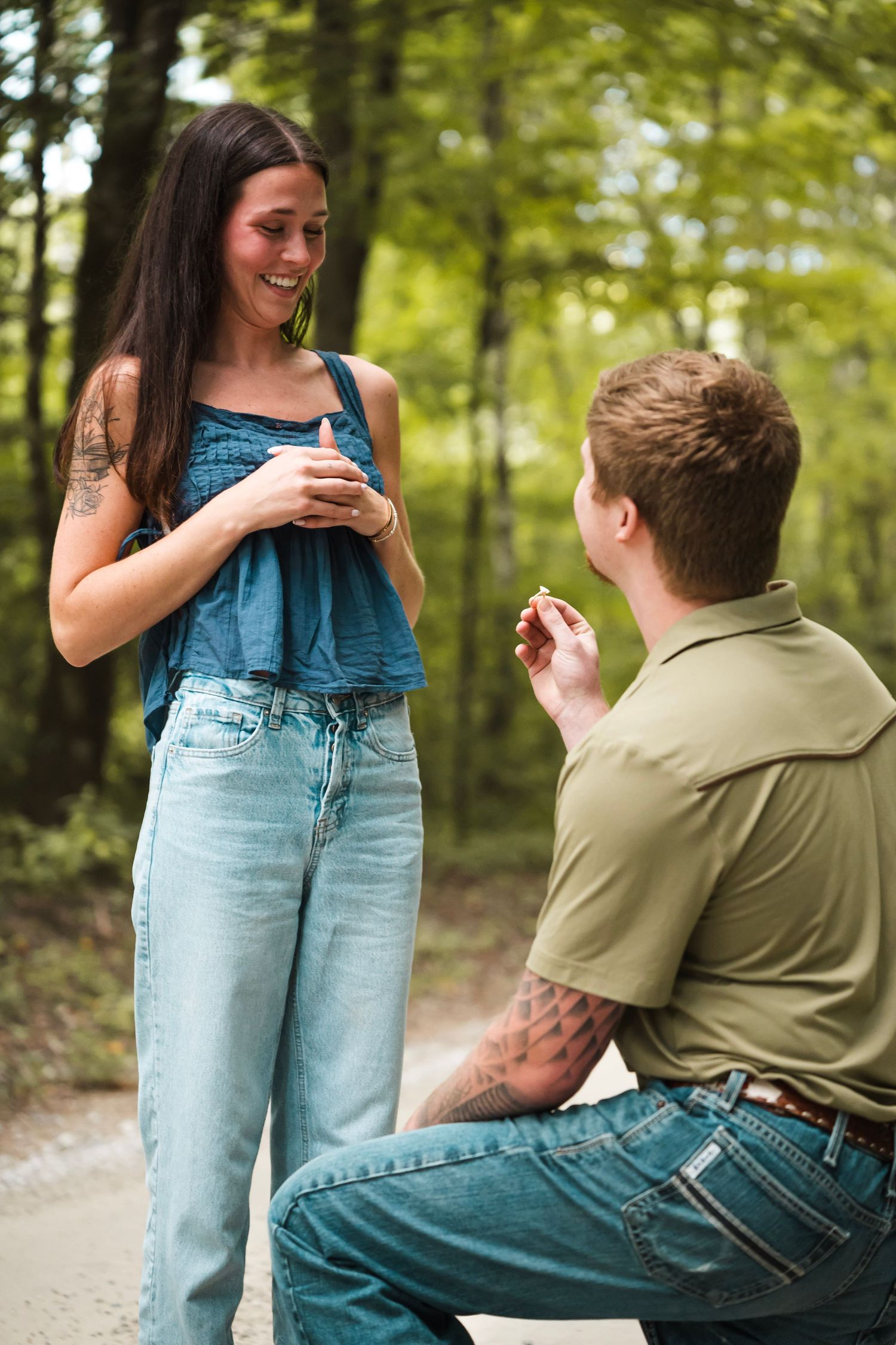 Man kneeling on one knee proposing to a smiling woman on a forest path, holding an engagement ring.