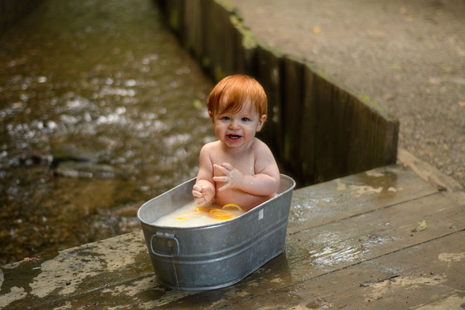 Happy red-haired baby sitting in a galvanized metal tub outdoors near a stream in a natural setting.