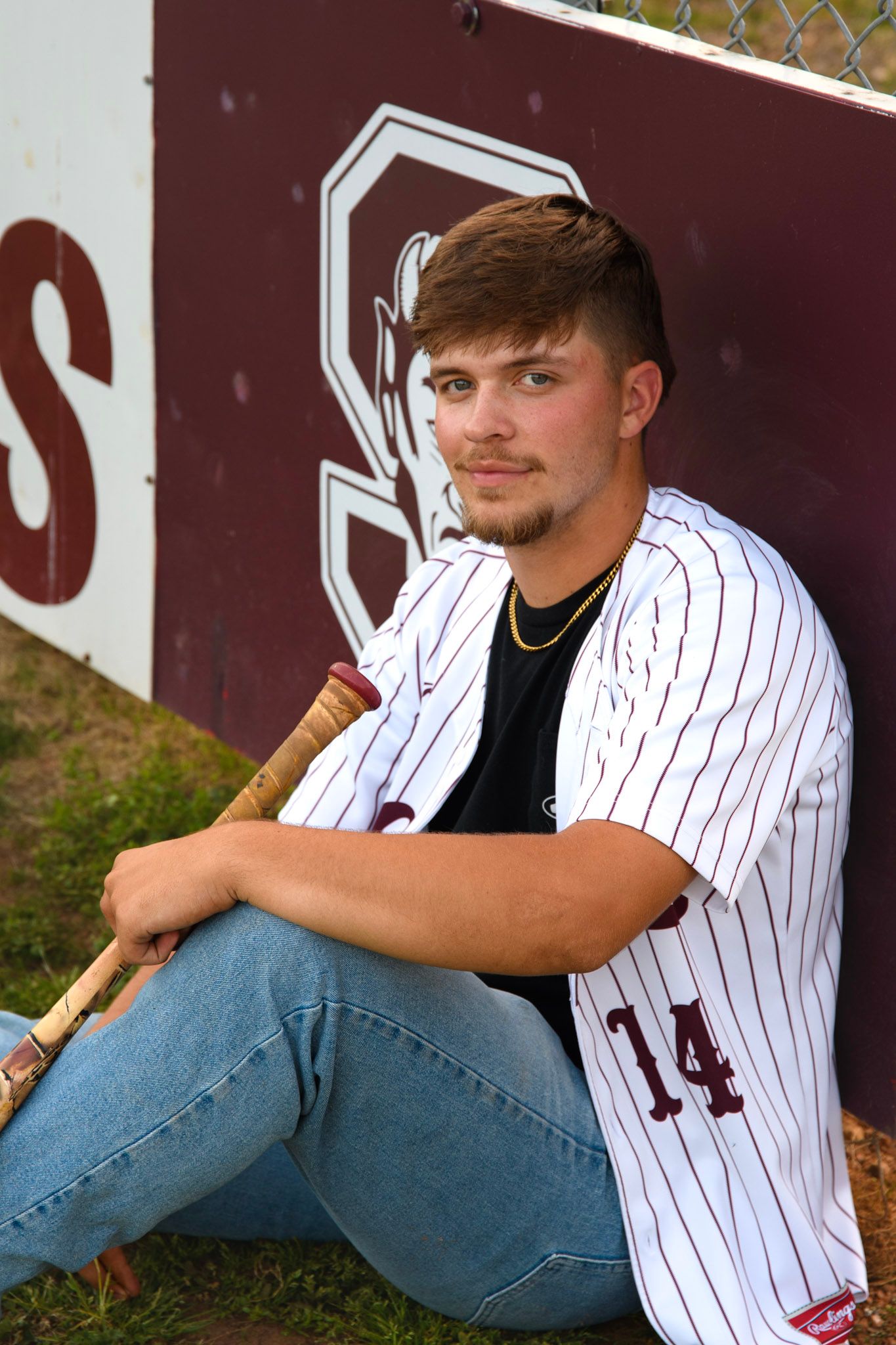 Young baseball player in jersey number 14 sits against dugout wall holding a bat during senior portrait session.