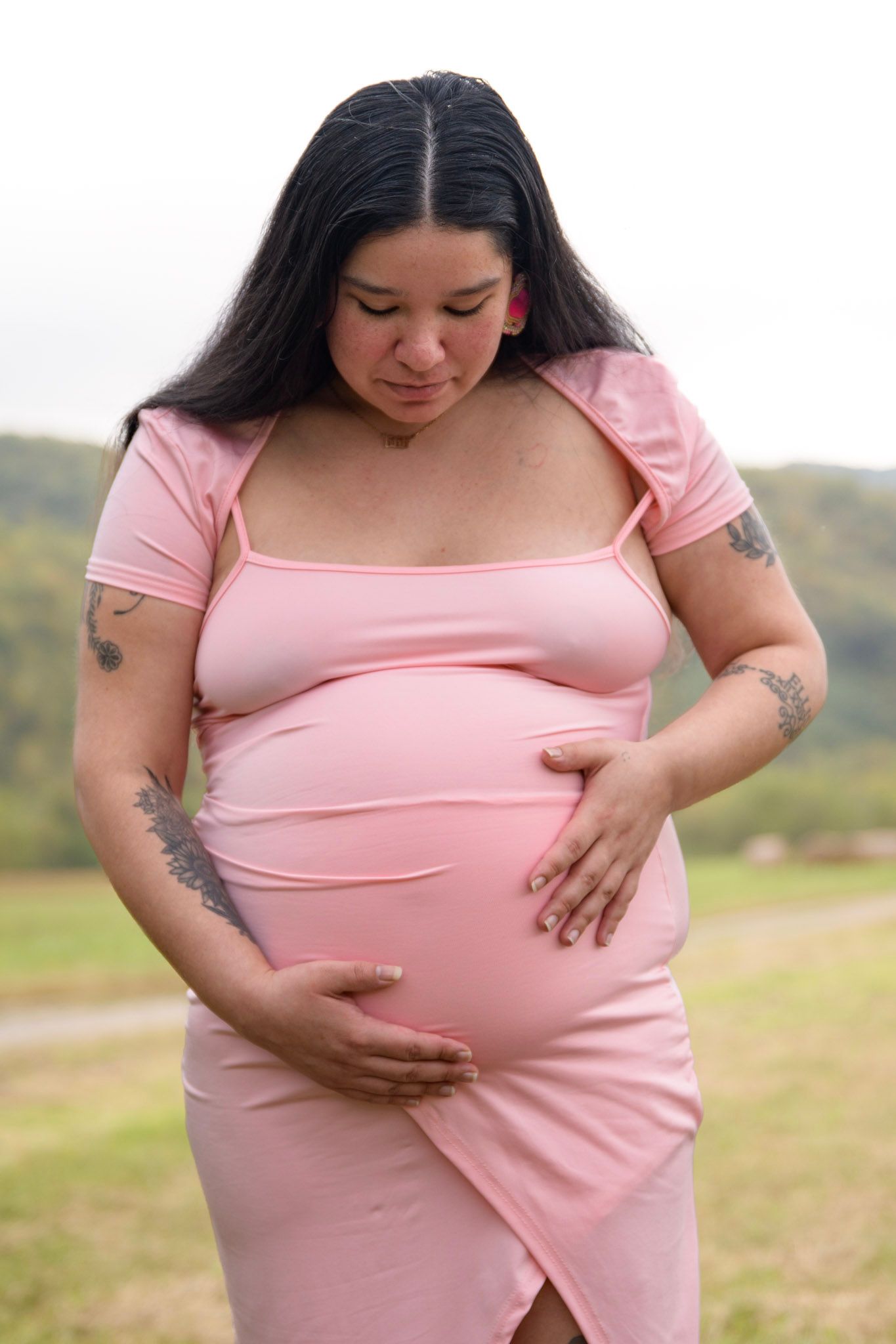Pregnant woman in pink dress cradling her baby bump while standing in an outdoor field setting.