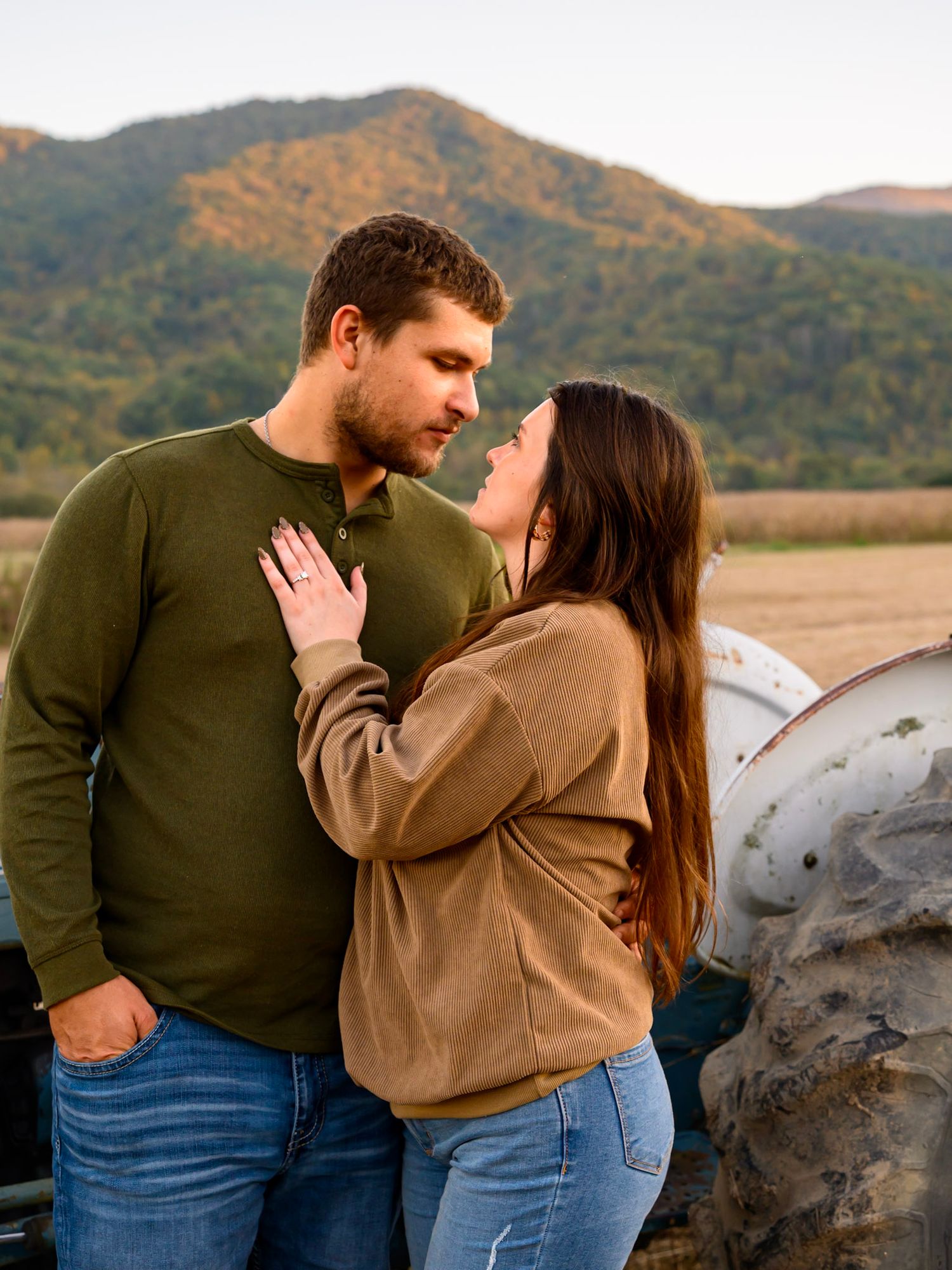Engaged couple embracing in front of a tractor with autumn mountains behind them, her ring hand resting on his chest.
