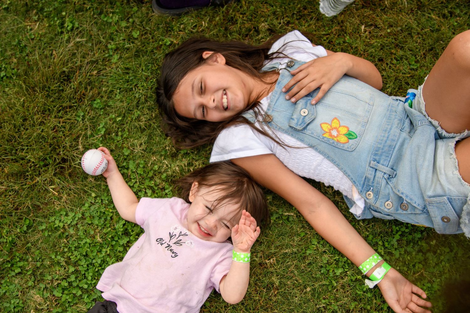 Two girls lying on green grass, older girl in denim vest smiling, younger toddler holding a baseball ball.