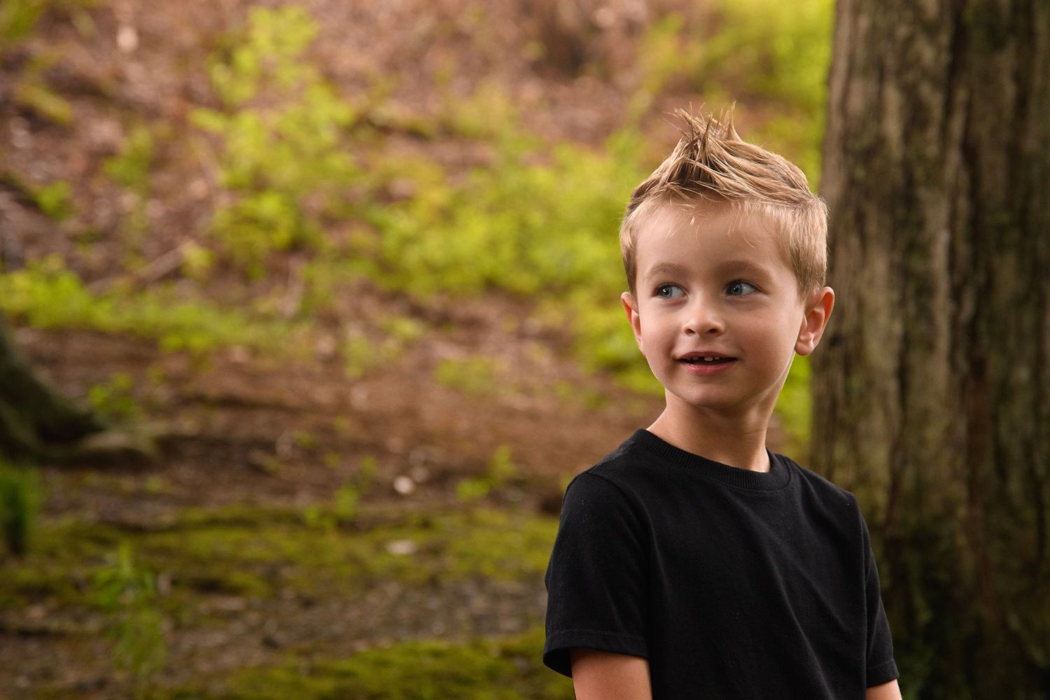 A young boy with blonde hair wearing a black t-shirt smiles outdoors near a moss-covered tree in a lush green forest.