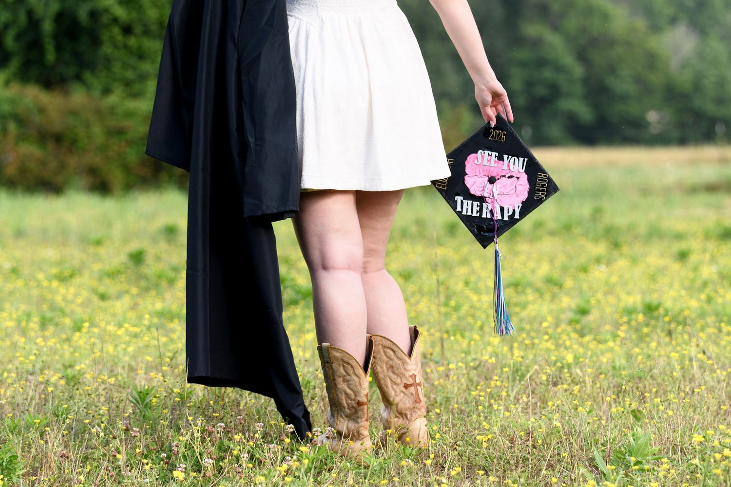 Graduate in white dress and cowboy boots holding decorated mortarboard cap in a sunny field.