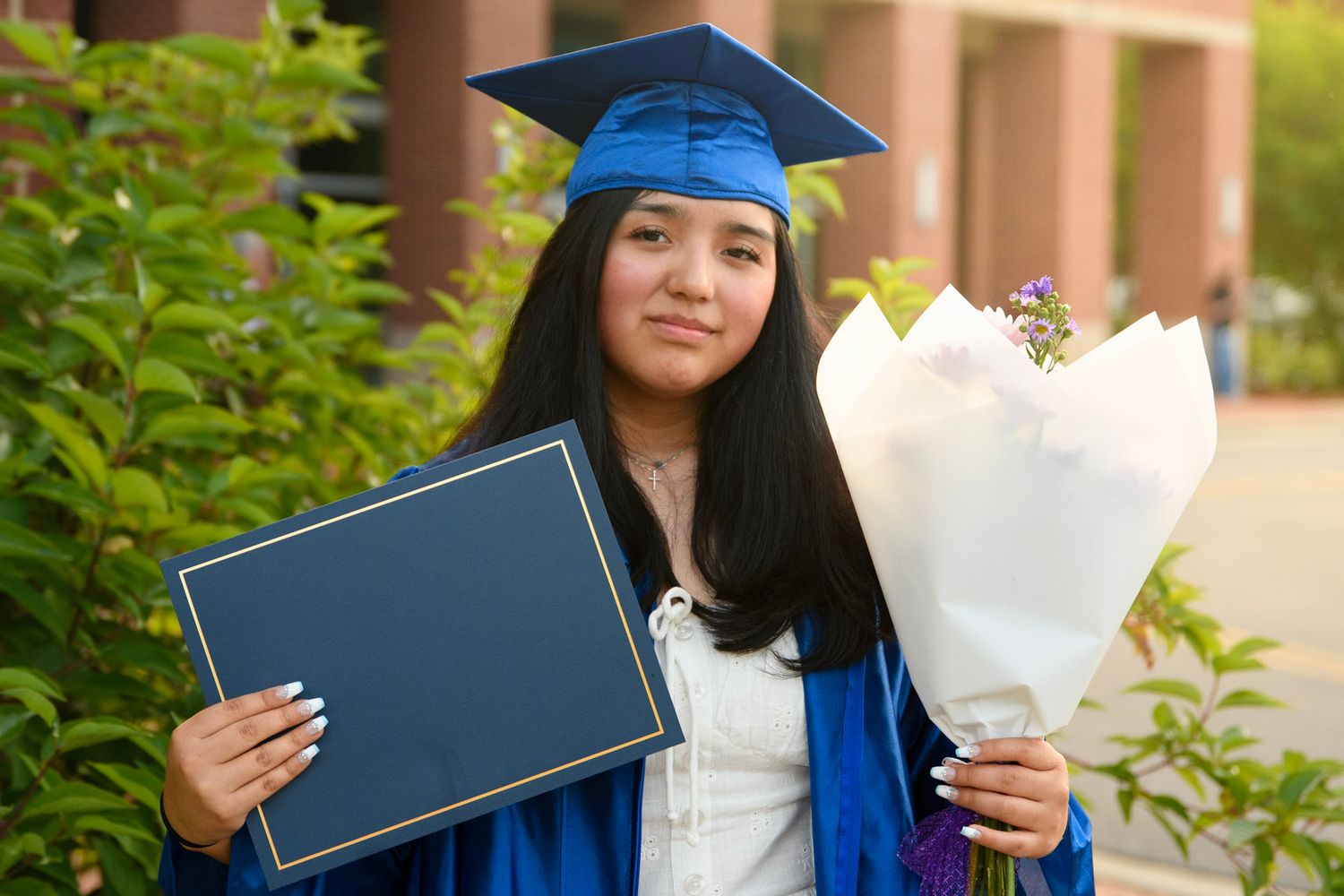 Young woman in blue graduation cap and gown holding diploma and flower bouquet outdoors.