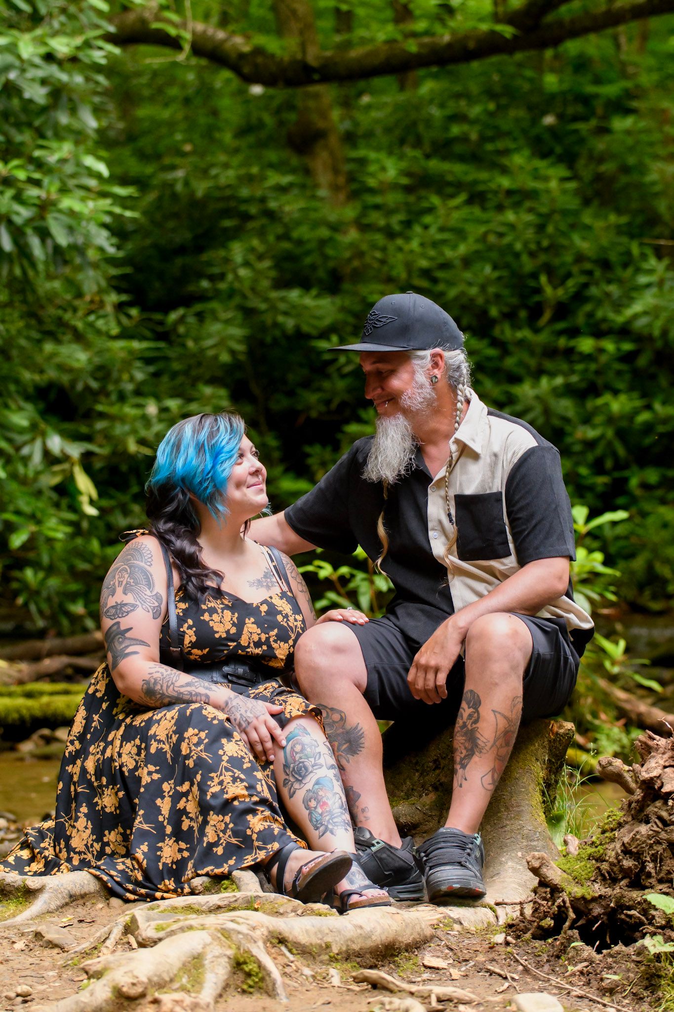 Tattooed couple with blue hair and beard sitting on rocks in lush green forest, smiling at each other.