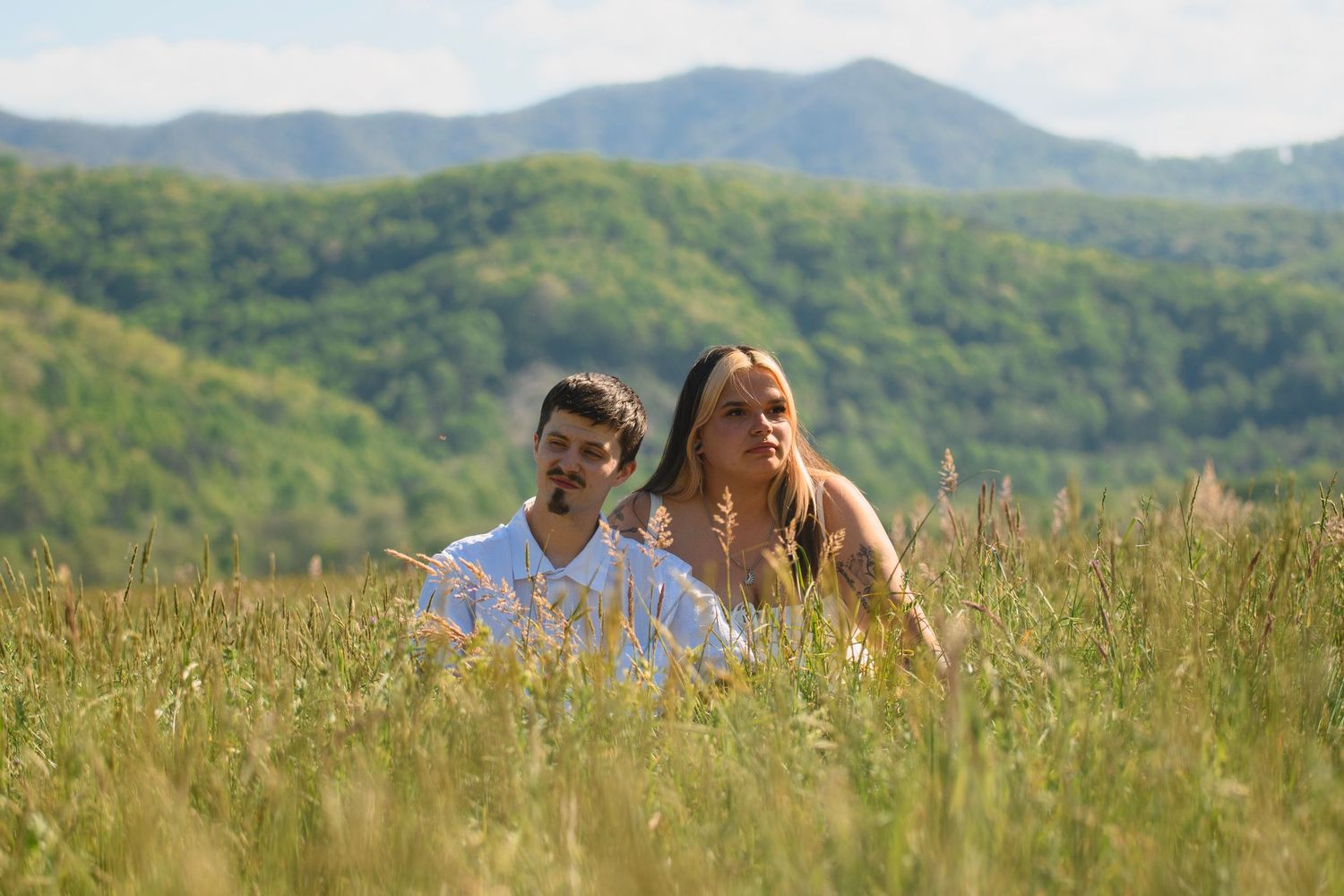 Young couple sitting together in tall grass field with green mountain backdrop on sunny day.
