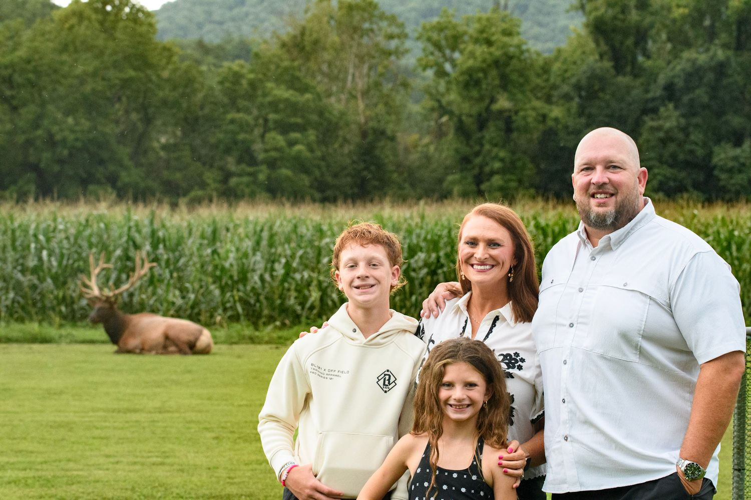 Family of four smiling outdoors near a cornfield with a deer resting in the background on a green lawn.
