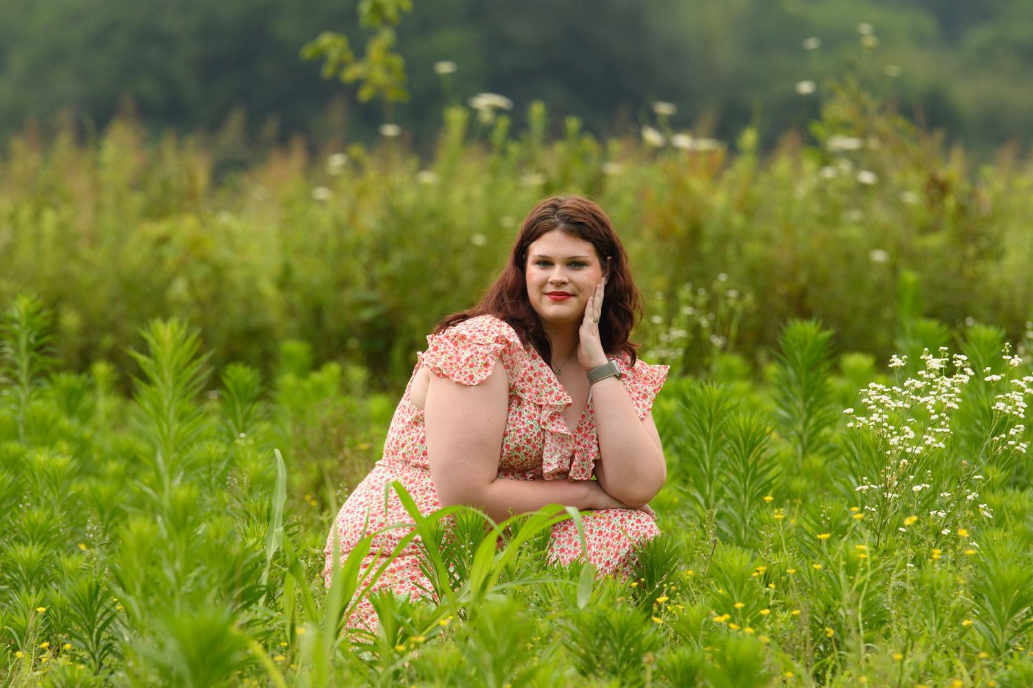 Young woman in floral dress sitting gracefully in a lush green meadow with wildflowers on a misty day.