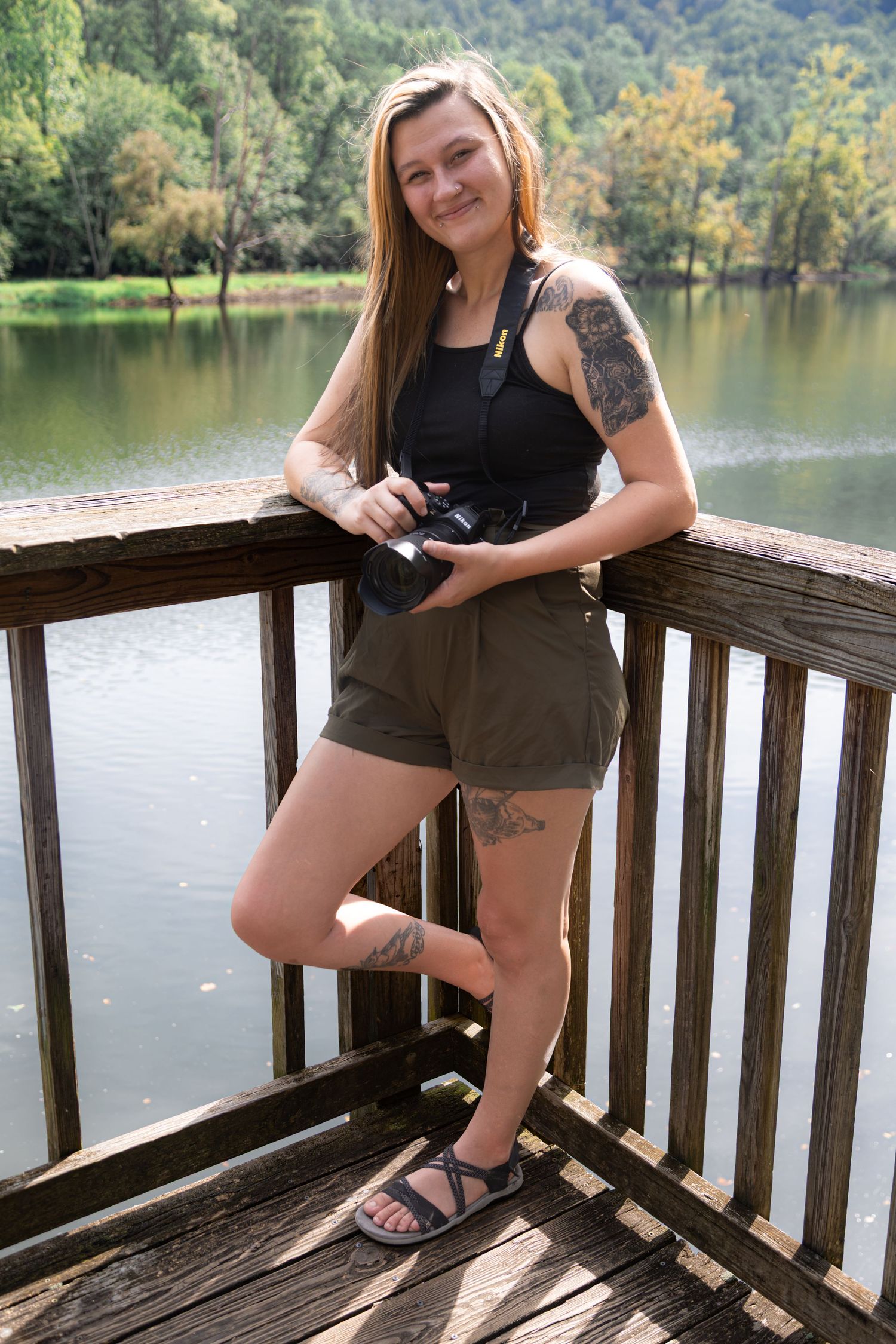 Young woman with tattoos holding a camera on a wooden dock overlooking a serene lake surrounded by trees.