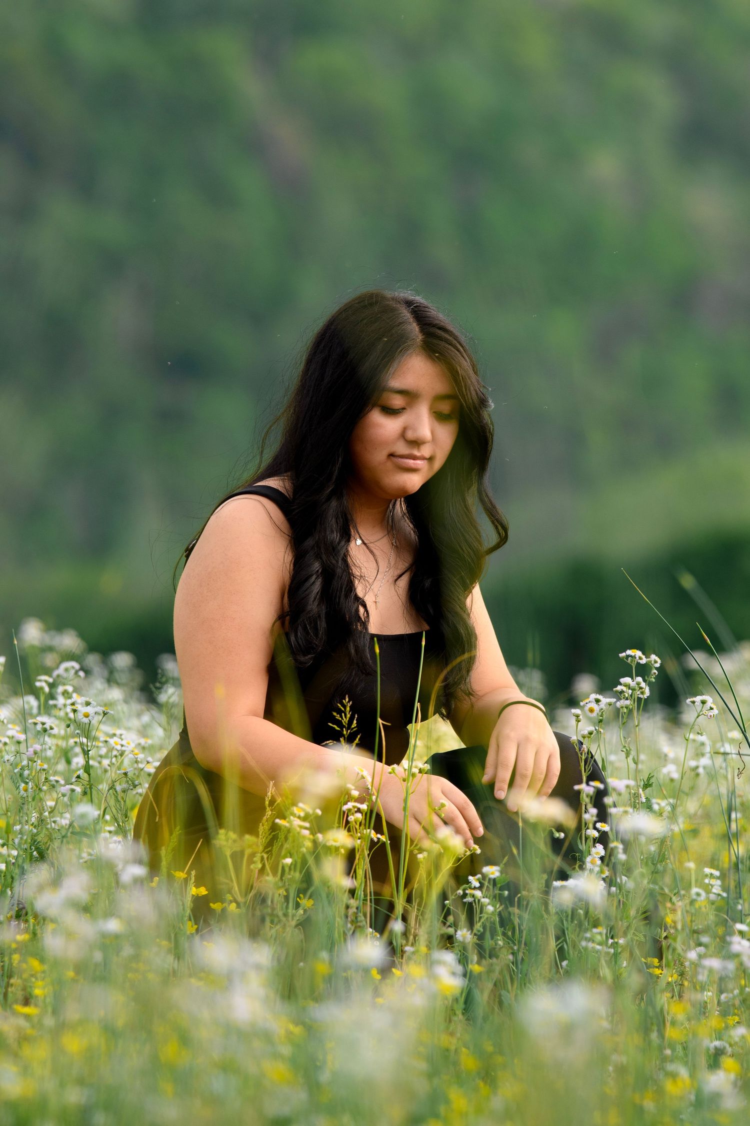 Young woman with long dark hair sitting among white wildflowers in a green meadow picking flowers.