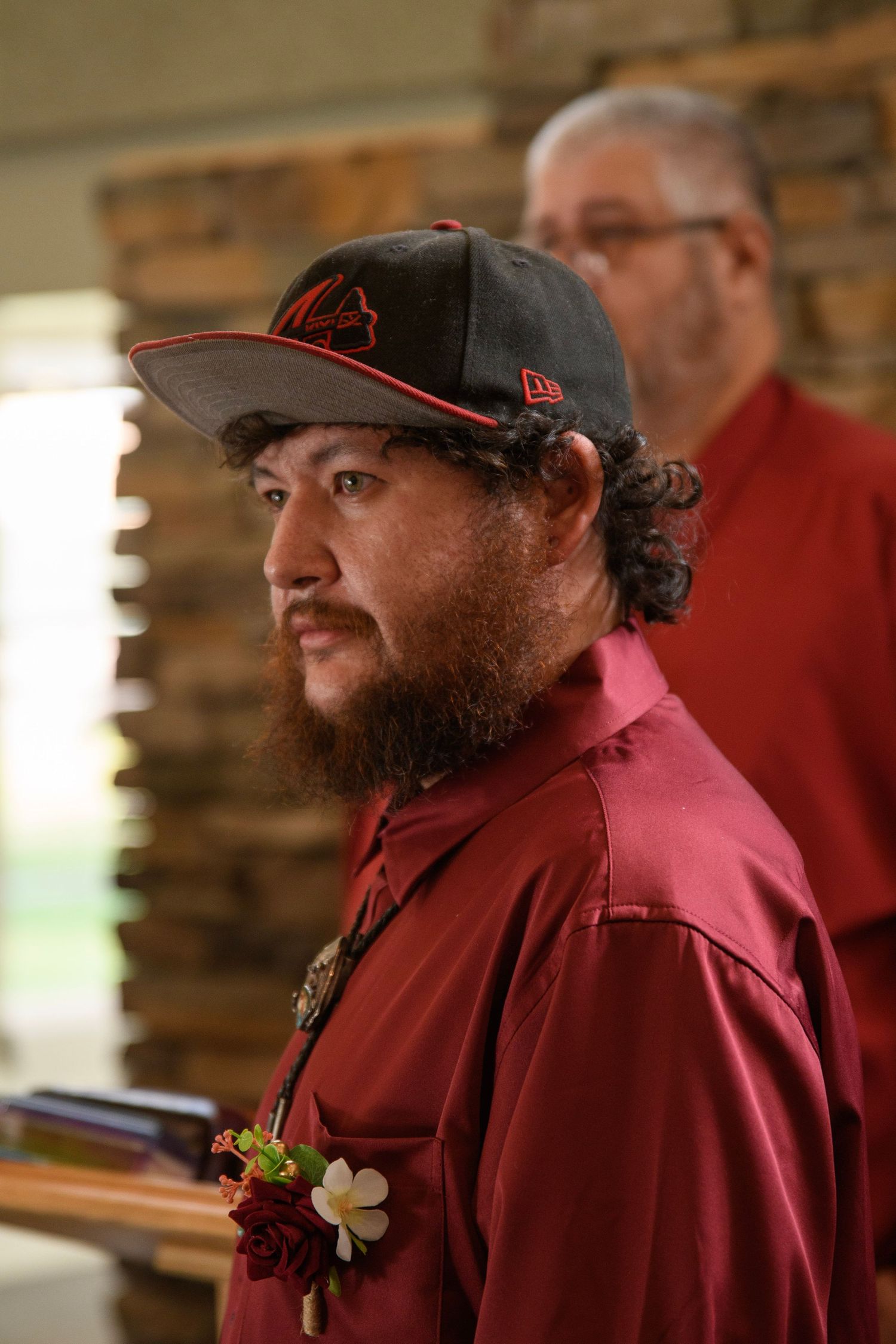 Bearded groom in red shirt and black New Era cap with floral boutonniere at indoor wedding ceremony.