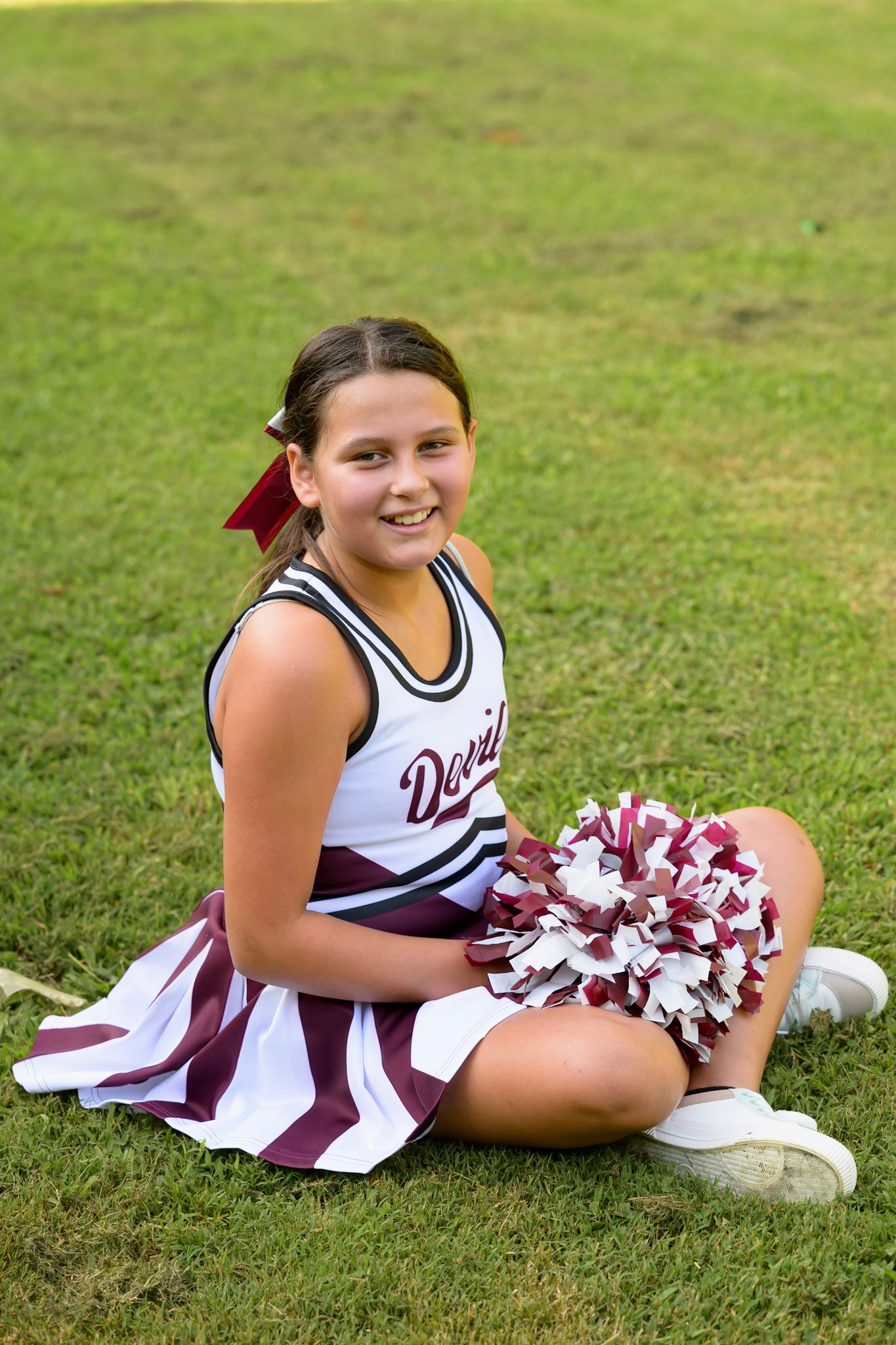 Smiling young cheerleader in Devils uniform sitting on grass holding maroon and white pom-poms.