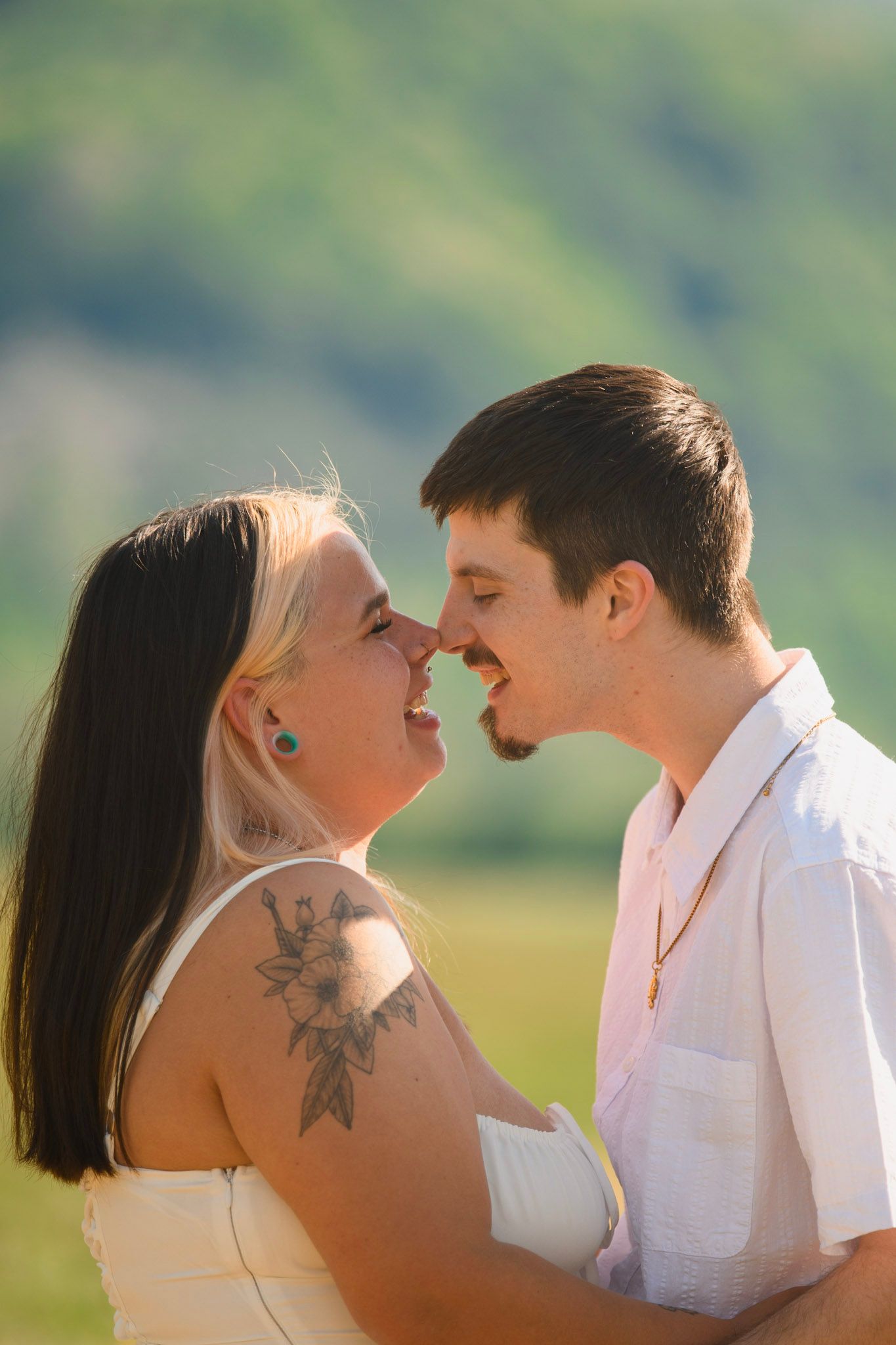 A couple shares a romantic nose-to-nose moment outdoors, with the woman showing a floral arm tattoo and blonde highlights.