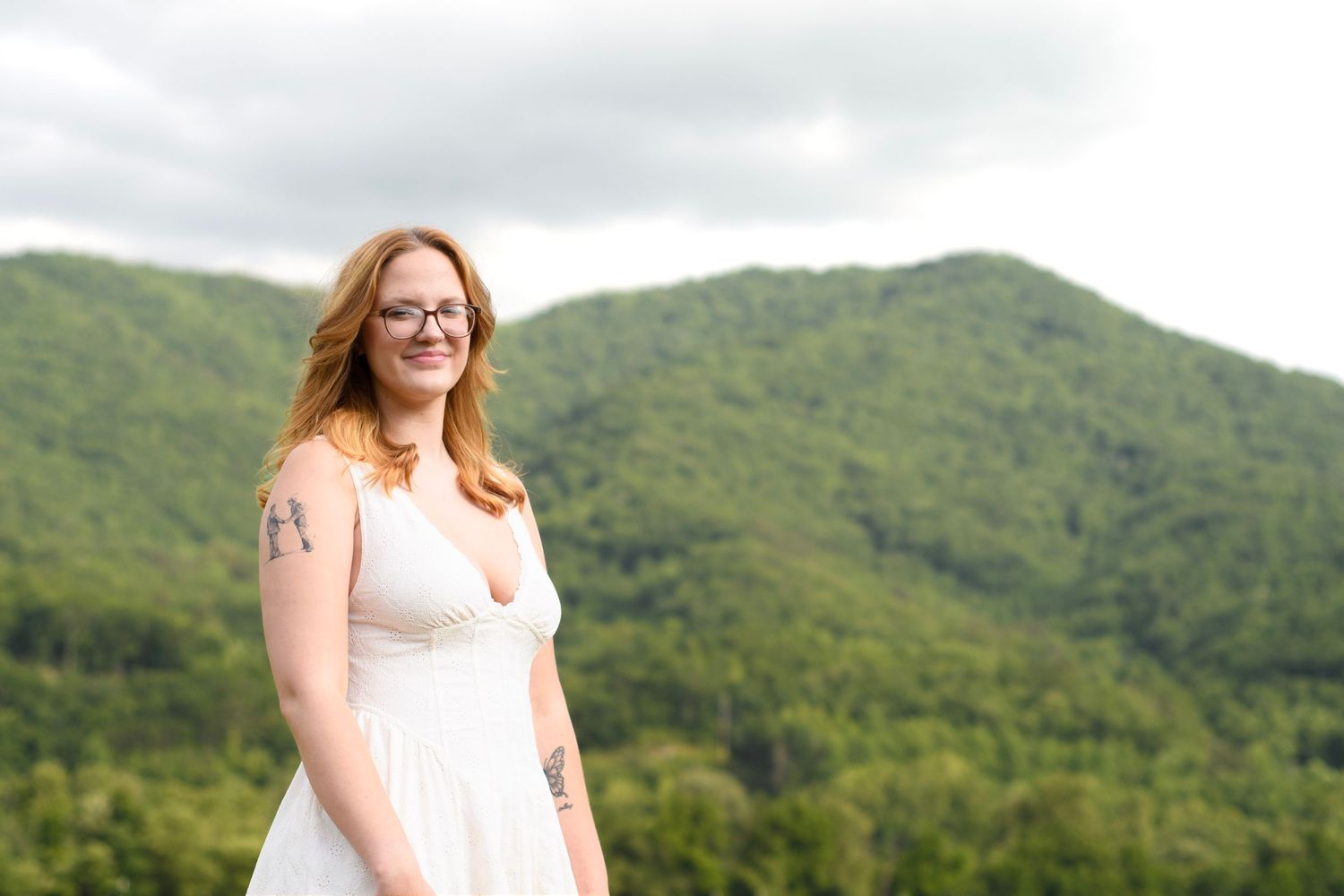 Smiling redhead woman in white dress with tattoos poses before lush green mountain landscape.