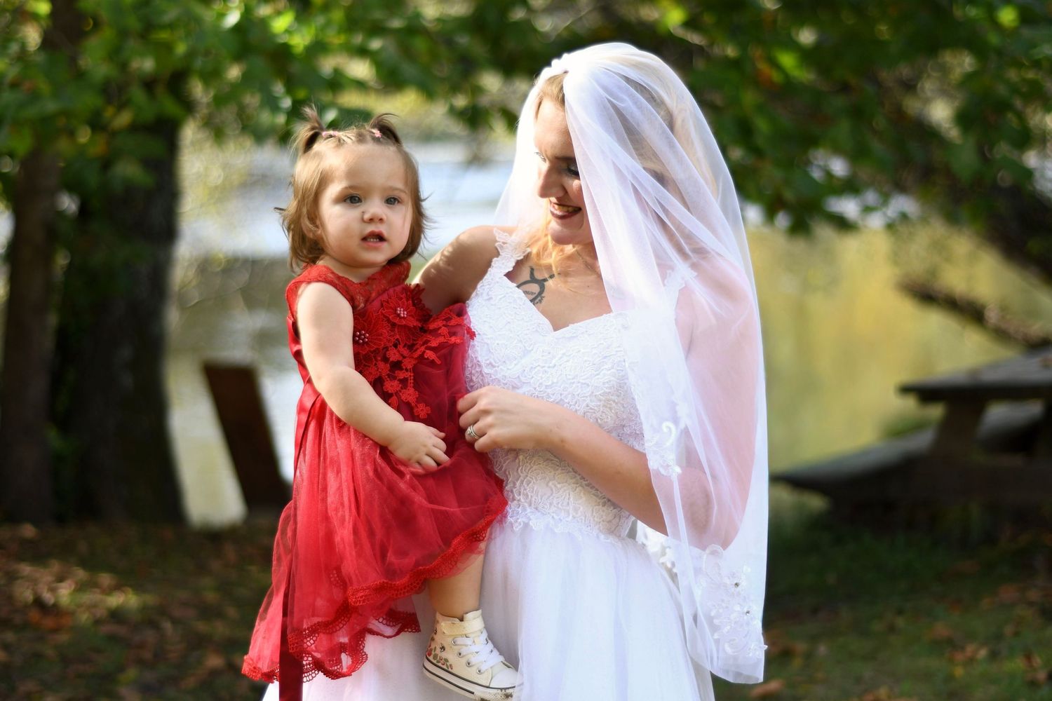 Bride in white wedding gown and veil holding young girl in red dress outdoors near trees.