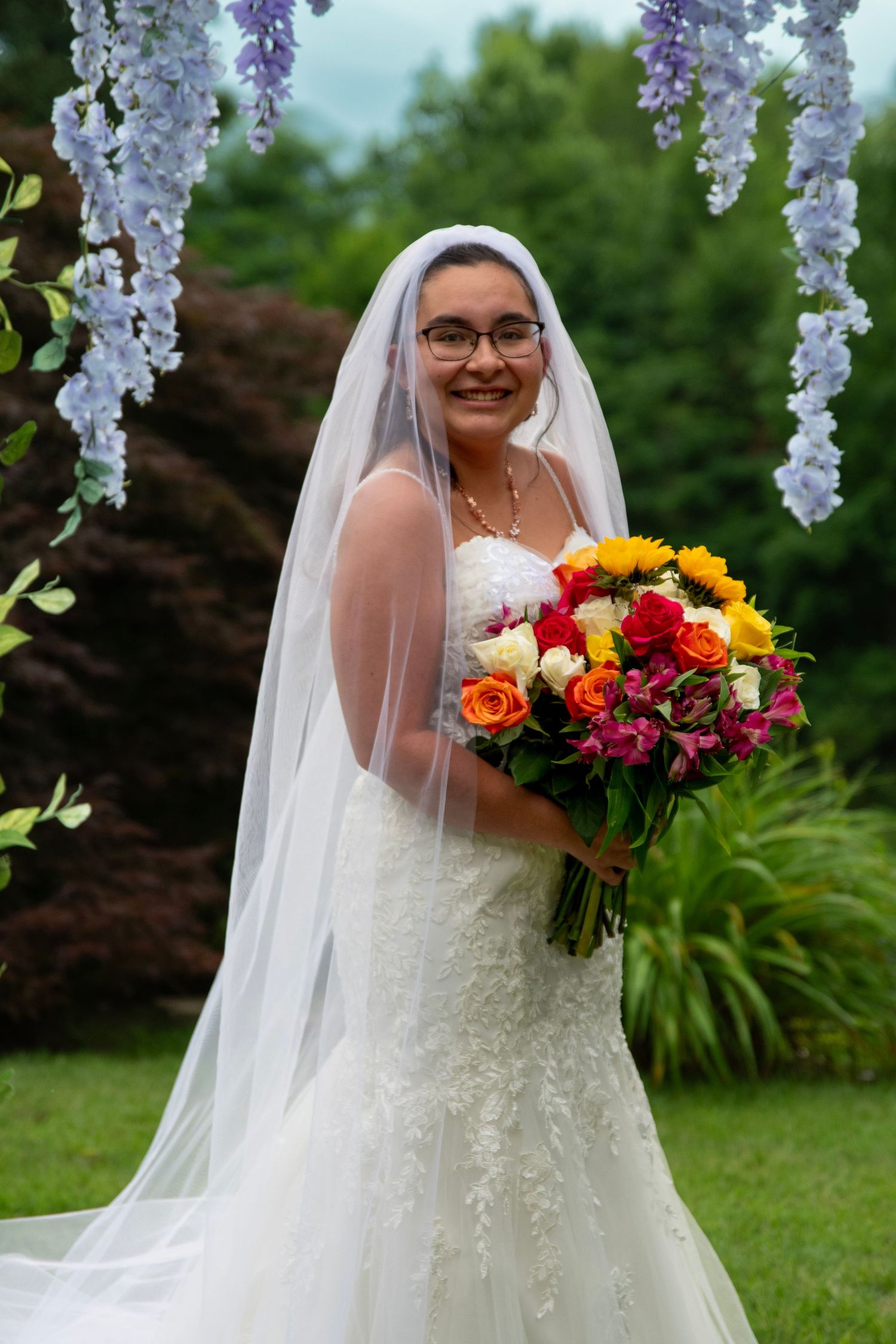 Smiling bride in white lace mermaid gown holding colorful bouquet under purple wisteria at outdoor wedding.
