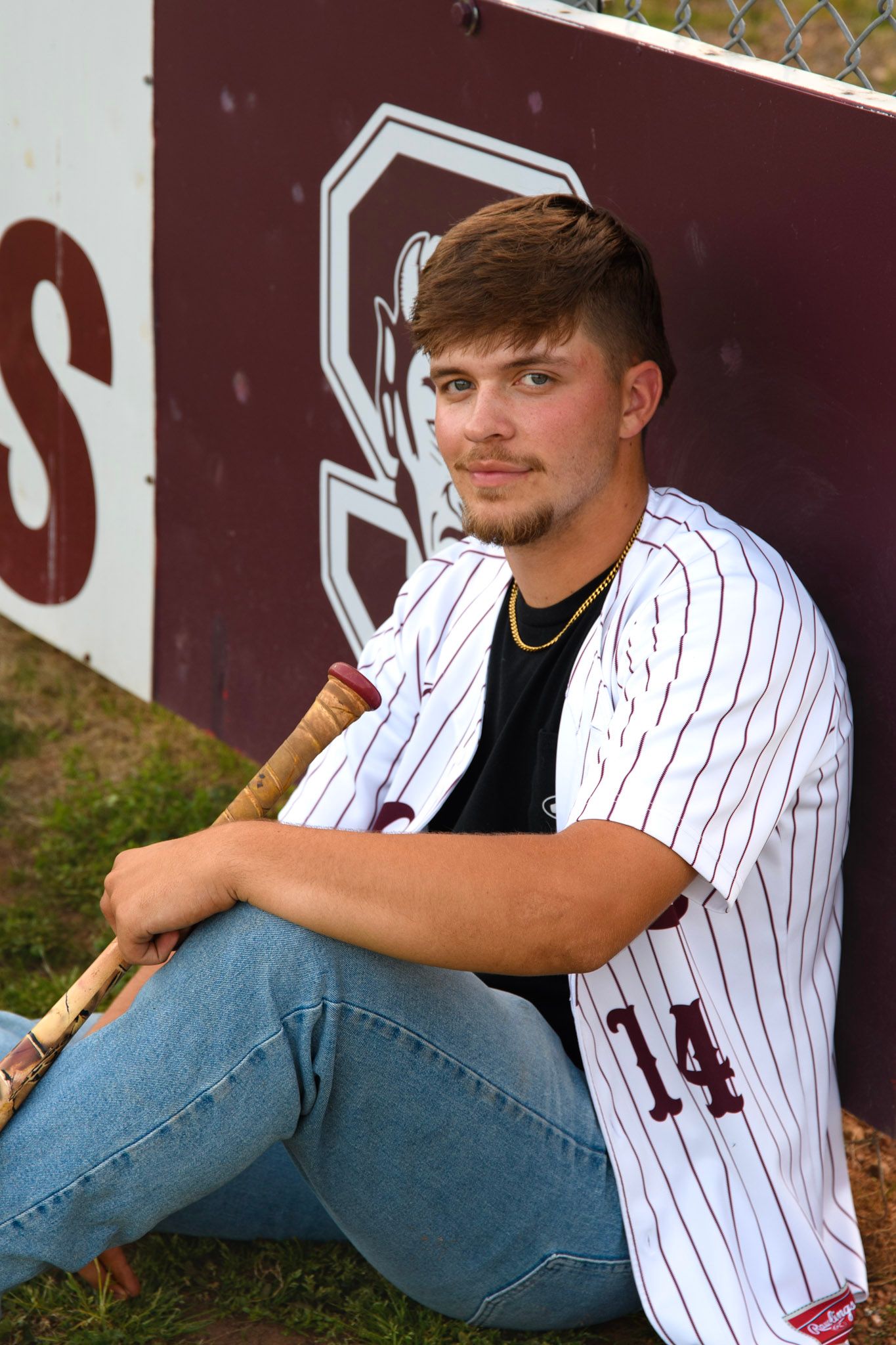 Male baseball player in maroon jersey number 14 sitting against a dugout wall holding a bat.