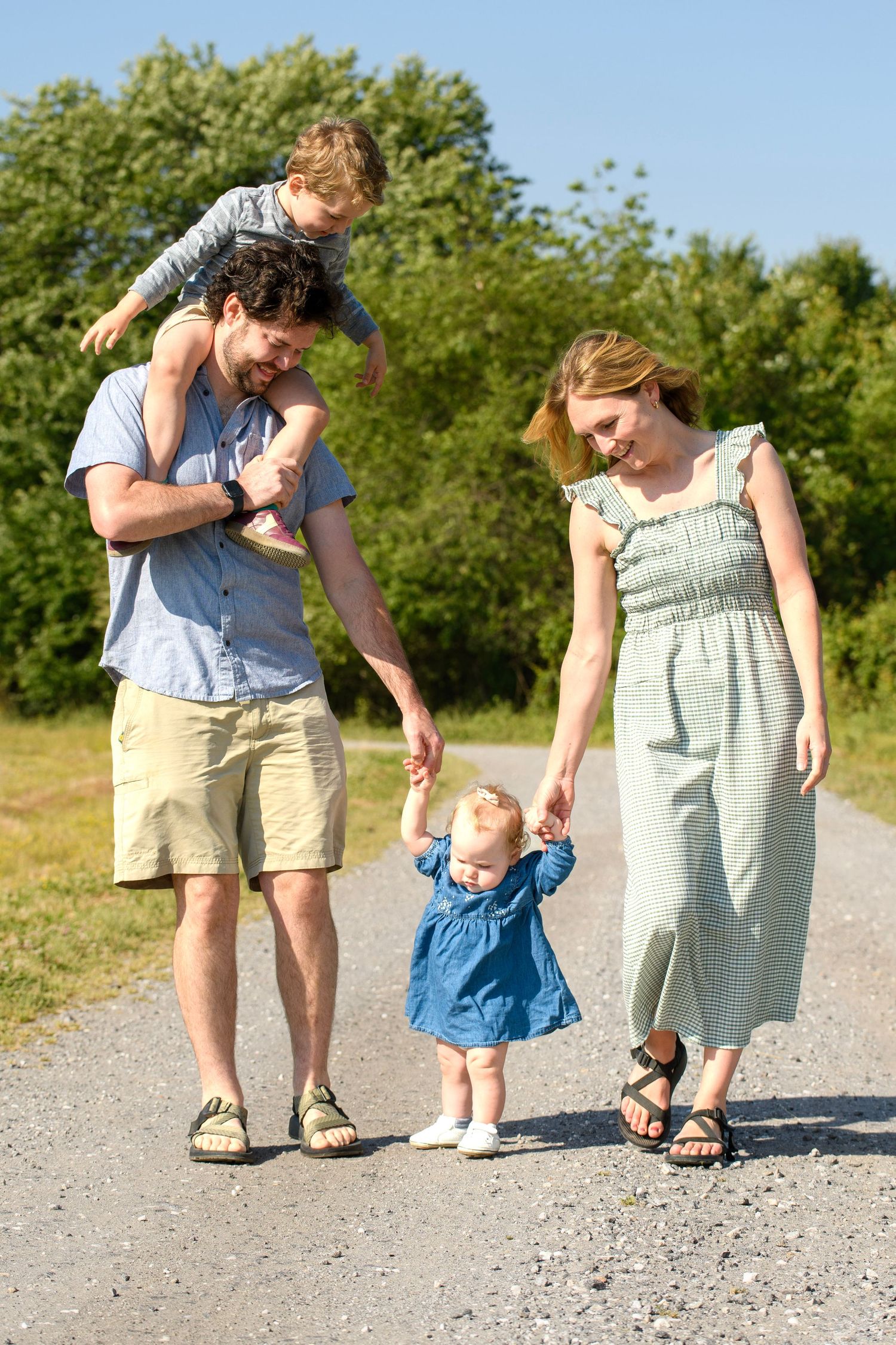 Family of four walking on a gravel path, dad carrying child on shoulders outdoors.