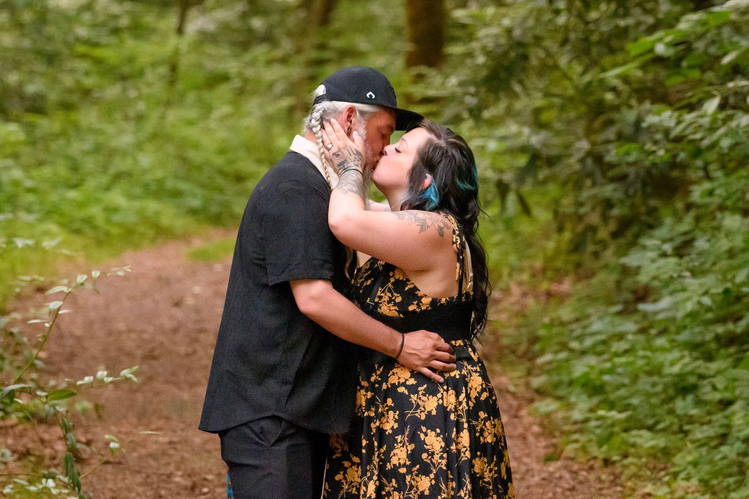 Couple sharing a romantic kiss on a forest trail, woman in floral dress, man in black outfit, surrounded by lush greenery.